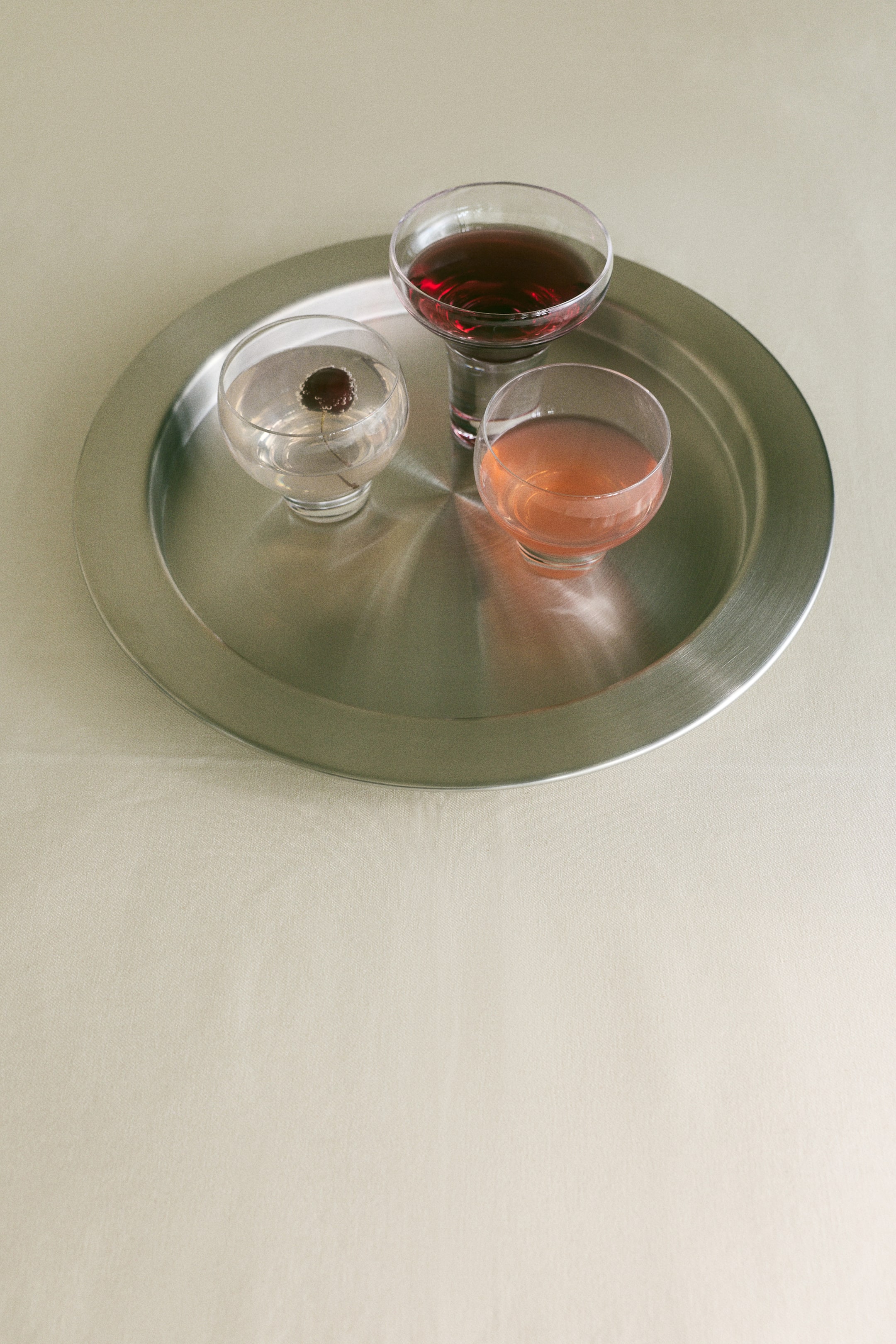 Grotere afbeelding bekijken: Drie mondgeblazen glazen tumblers met stevige bodems staan op een ronde, geborstelde metalen dienblad. De tumblers, met afgeronde kelken en dikke, massieve glazen bodems, bevatten heldere, roze en donkerrode vloeistoffen.