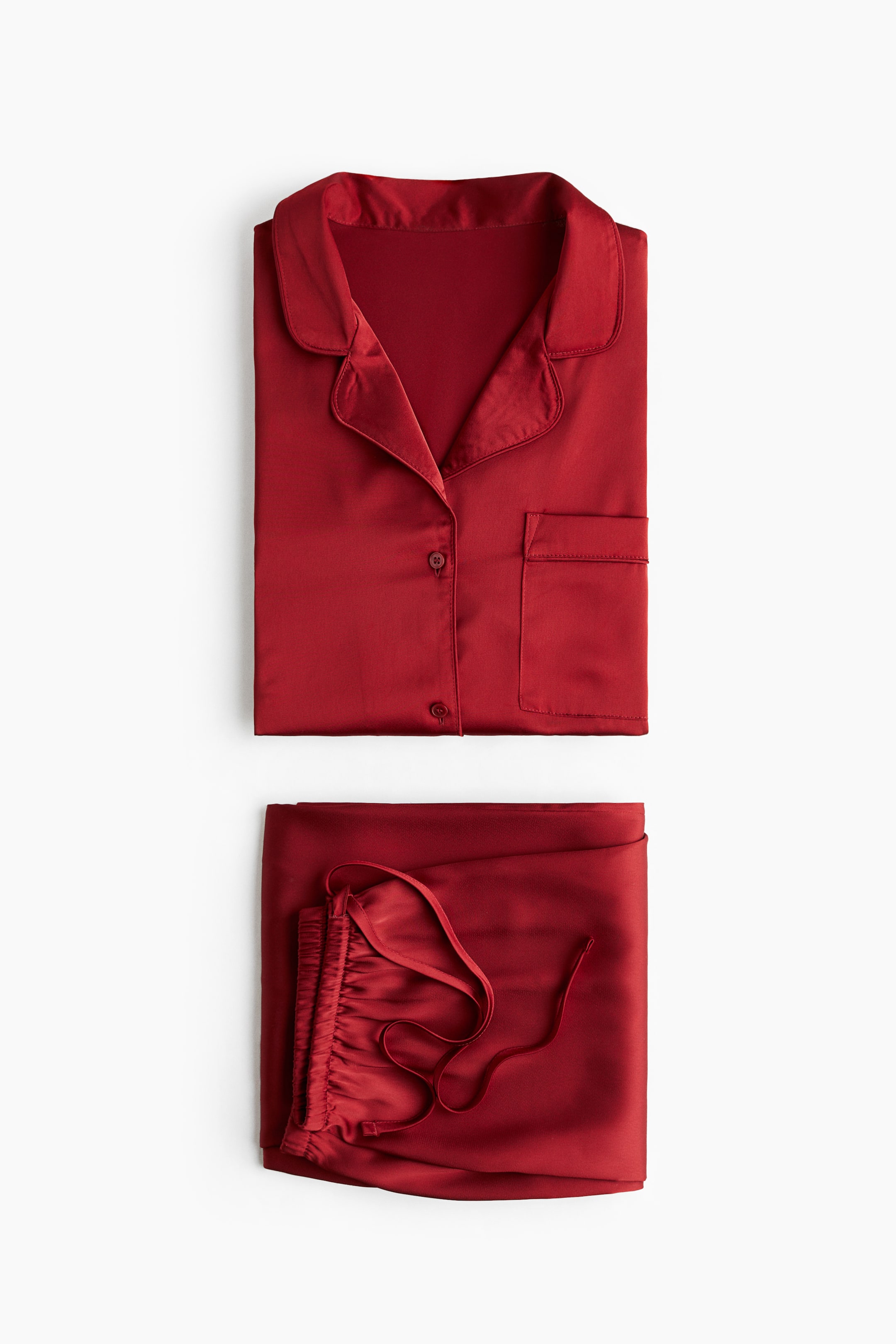 View larger image: A red satin pajama set is displaying, folded flat. The shirt is featuring a resort collar, buttons down the front, and an open chest pocket, while the matching bottoms are showing an elasticated, drawstring waist.