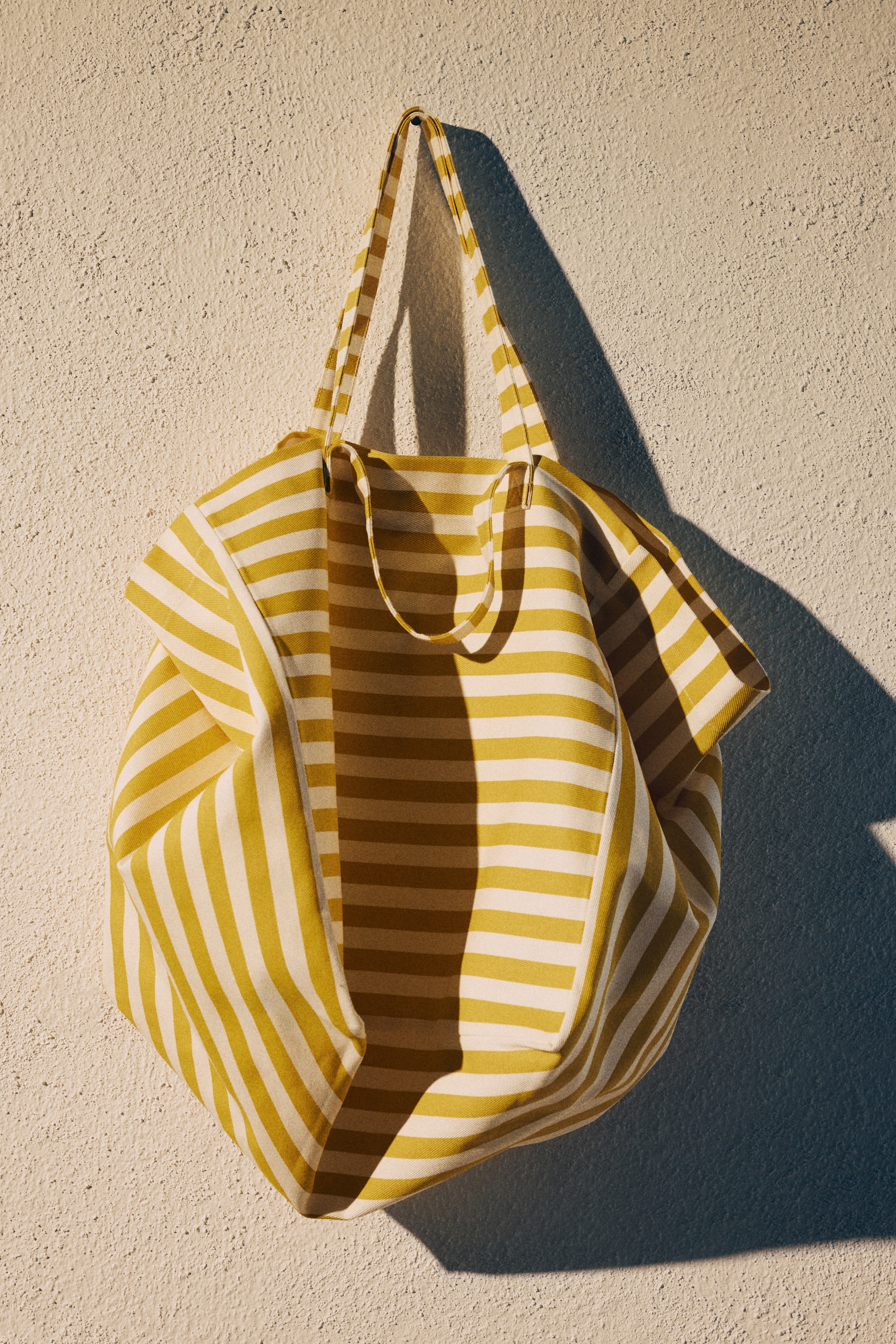 View larger image: A roomy striped cotton canvas beach bag is displayed from the front, showcasing wide yellow and off-white horizontal stripes. Two sets of handles are visible at the top, while the bag's upper edges fold inward, creating a relaxed, voluminous shape.