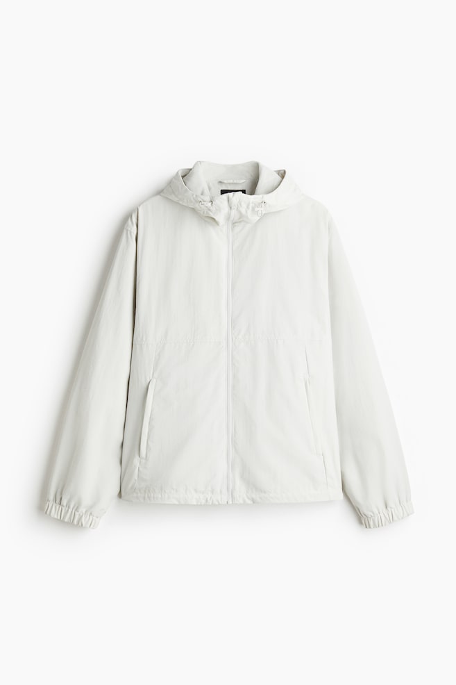 A light-colored windbreaker is presenting its FRONT, featuring a hood with a visible drawstring, a full zip closure, long sleeves with elasticated cuffs, two vertical zipped side pockets on the front, and an elasticated hem. - 1