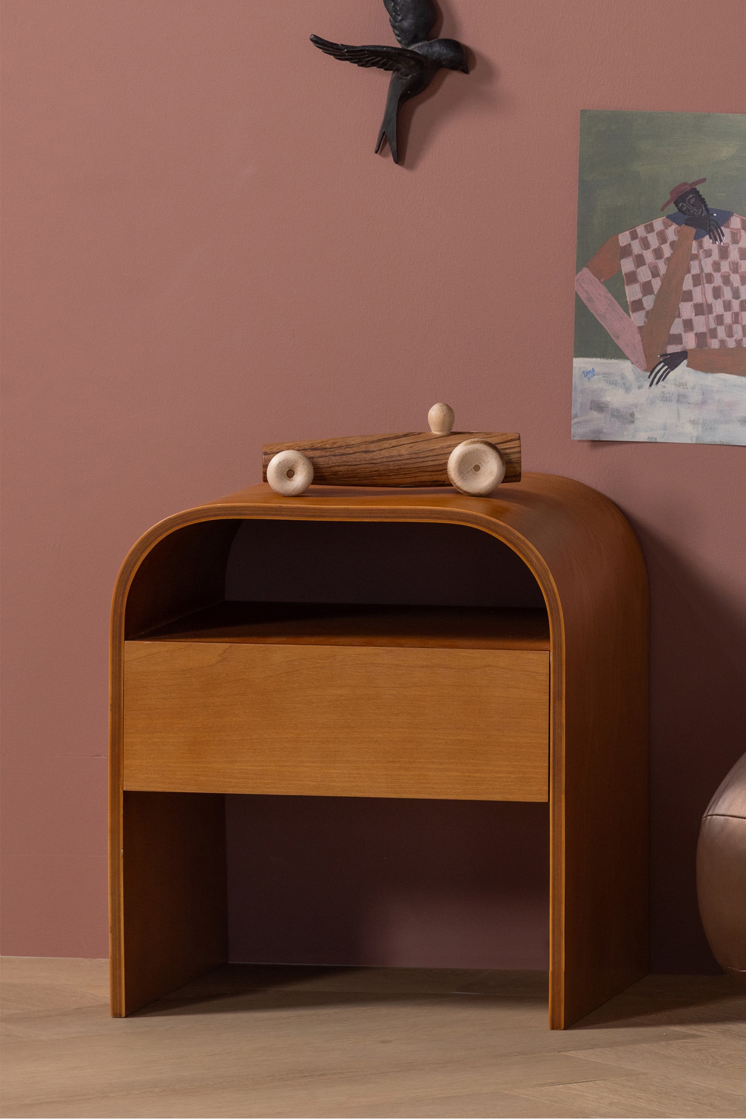 Een houten bijzettafel, lichtjes naar rechts gekanteld, toont een doorlopend gebogen bovenkant en zijkanten. Het beschikt over een open plank en een vlakke, greeploze lade die eronder is geïntegreerd, vervaardigd uit middelbruin hout met een zichtbare nerf. - 1