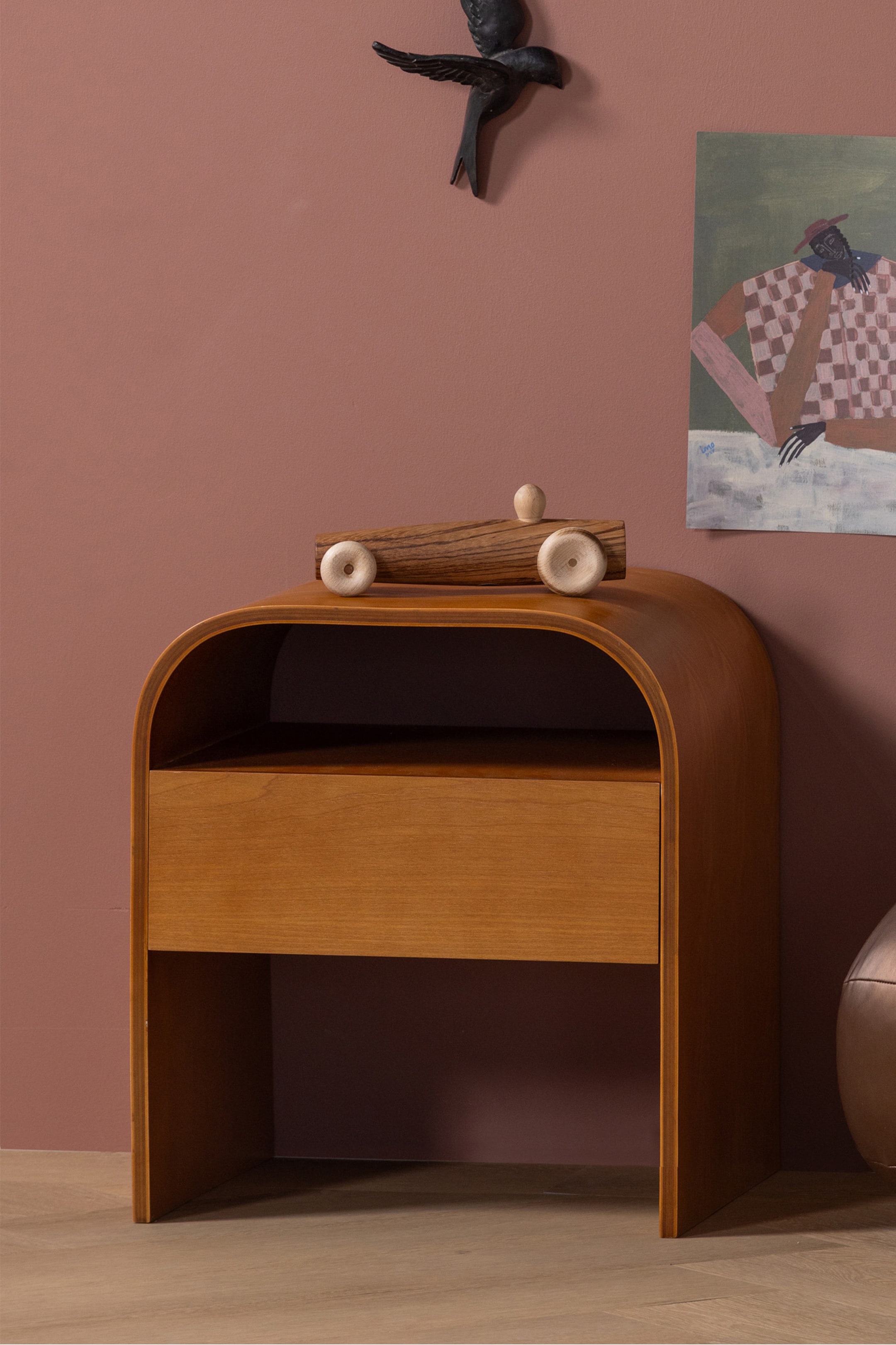 Grotere afbeelding bekijken: Een houten bijzettafel met een gebogen ontwerp, een open bovenste plank en een enkele lade eronder, gezien vanuit een lichte hoek. Een houten speelgoedauto rust op het bovenoppervlak.