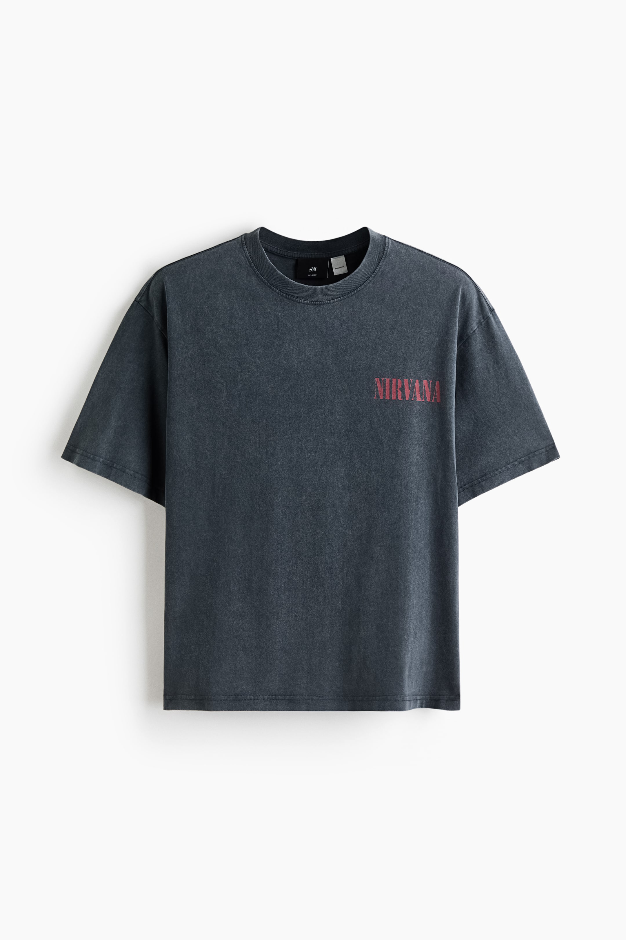 Agrandir l'image: Un t-shirt gris foncé est suspendu, arborant un délavage estompé et un col rond. Il présente des manches courtes et le mot 'NIRVANA' imprimé en rouge vieilli sur le haut de la poitrine gauche.