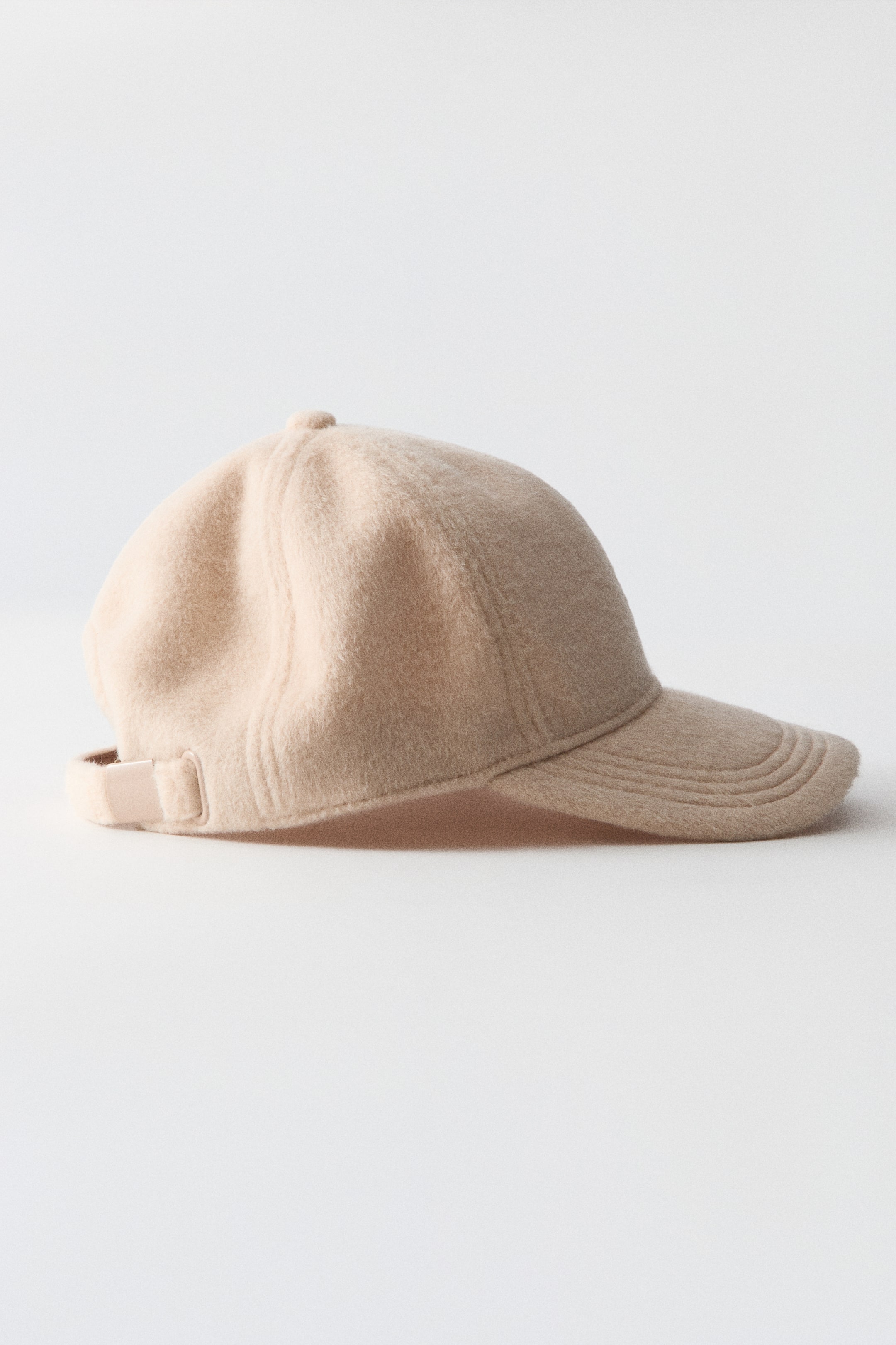 Ampliar la imagen: Una gorra beige de tela afelpada se encuentra en vista de perfil lateral, mostrando su superficie suave y texturizada, así como su construcción por paneles. Presenta una visera curva con detalles de costura y una tira ajustable con un broche metálico en la parte trasera.