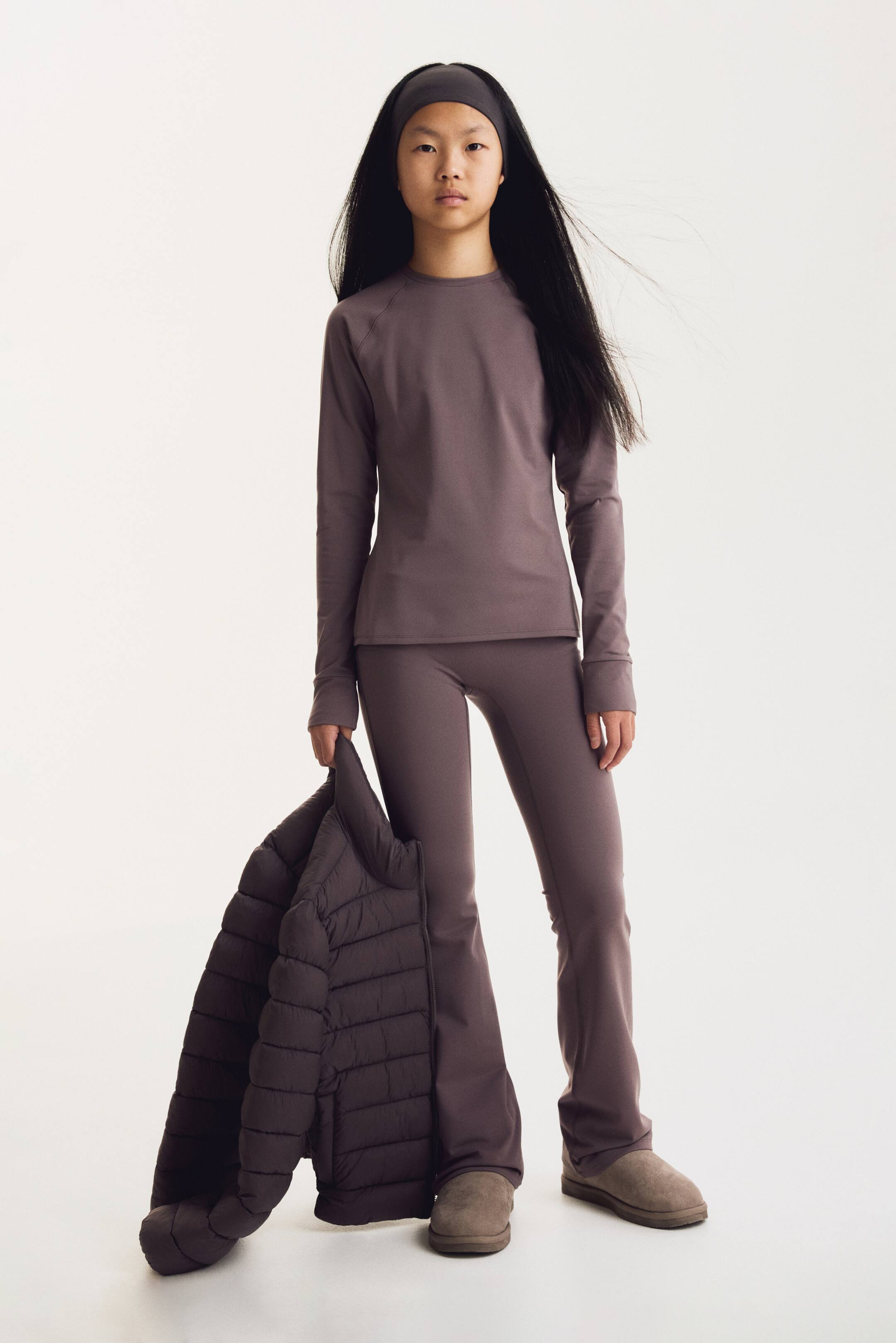 Visualizza l'immagine ingrandita: Un completo activewear coordinato in un delicato color malva è mostrato frontalmente, composto da un top a maniche lunghe con girocollo e pantaloni slim-fit svasati. La modella tiene in una mano una giacca puffer trapuntata marrone scuro, che cade verso il pavimento.