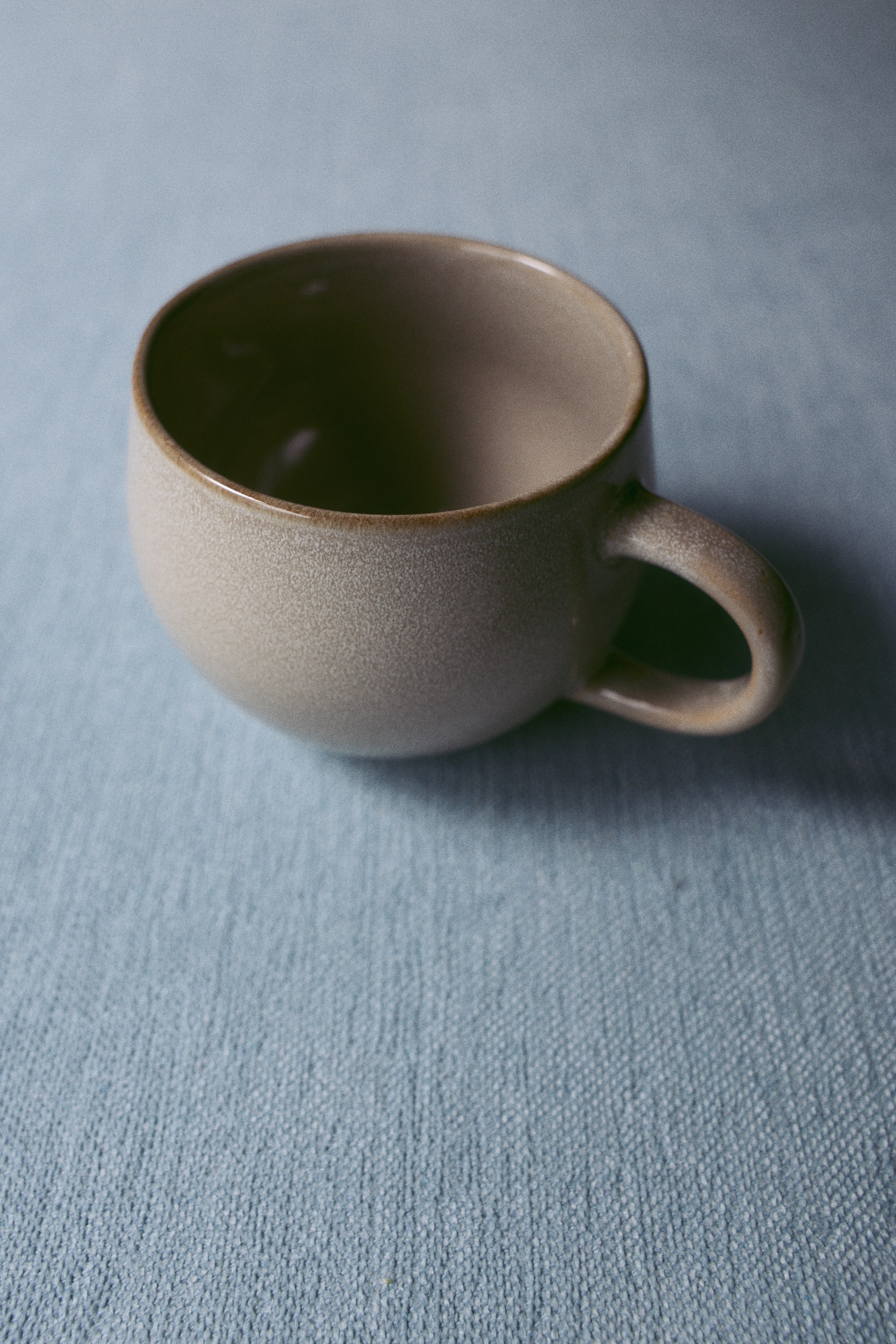 View larger image: A stoneware tea mug is shown with a rounded, wide-rimmed design and a speckled beige and light brown reactive glaze. The mug features a glossy brown interior, a slightly darker rim, and a curved handle on the right-hand side.