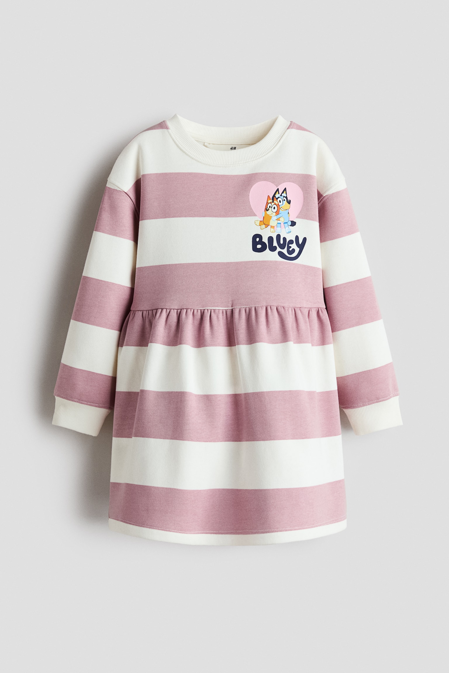 A long-sleeved sweatshirt dress is shown from the front, featuring wide horizontal stripes in dusty pink and off-white. The dress is designed with a ribbed crew neck, ribbed cuffs, and a gathered waist seam, with a graphic print on the upper left chest depicting the characters Bluey and Bingo inside a pink heart above the text "BLUEY."