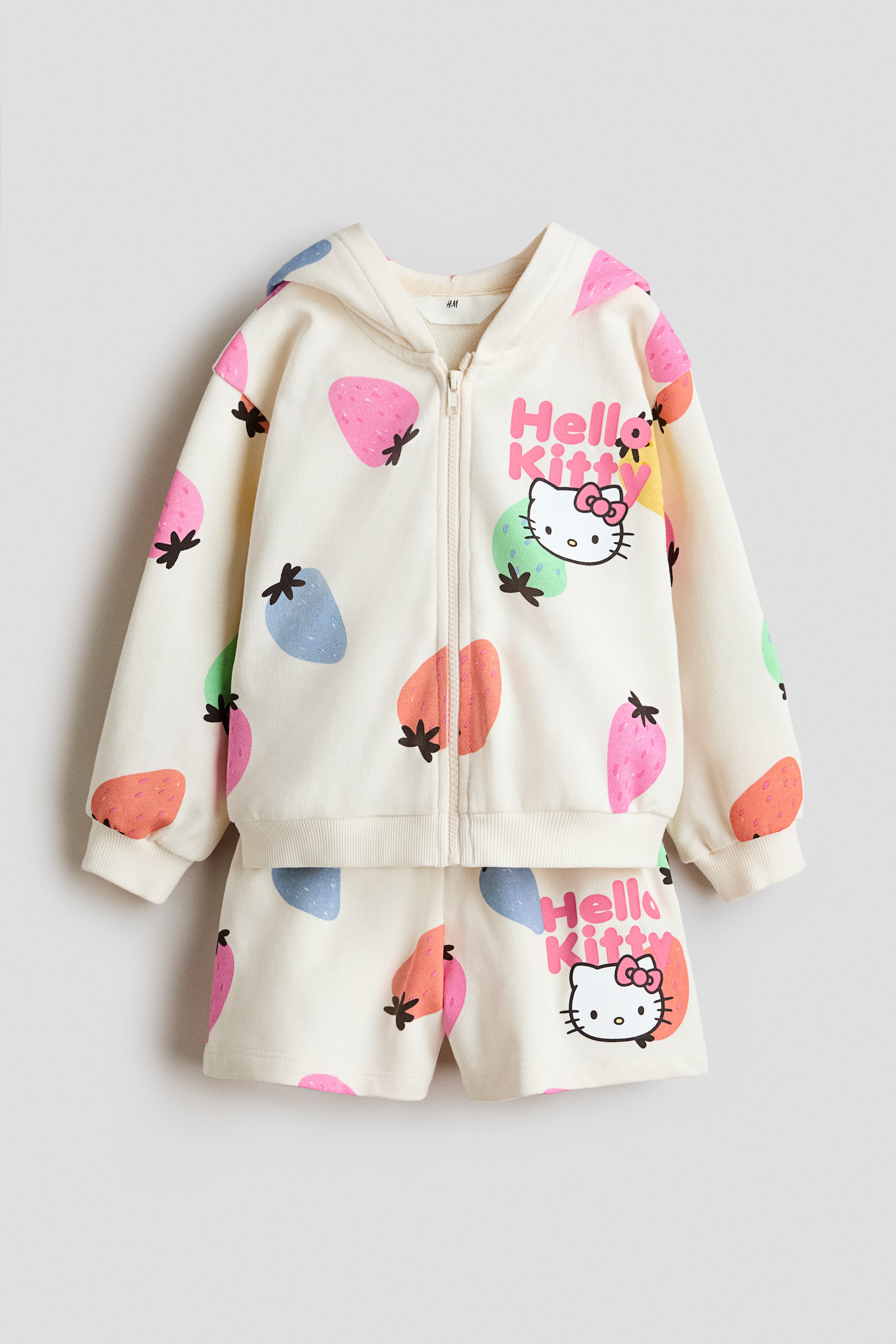 查看大圖: 奶油色套裝正面展示，包括拉鍊連帽衫和輕量印花運動衫面料的配套短褲。這套裝飾有色彩繽紛的莓果形狀和Hello Kitty圖案，連帽衫配有內襯帽兜、全拉鍊設計、落肩、長袖帶羅紋袖口和羅紋下擺，短褲則配有鬆緊腰帶。