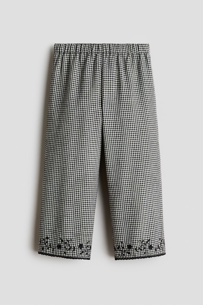 Estos pantalones de algodón sarga con estampado de pata de gallo en blanco y negro se muestran de frente, con una pretina elástica fruncida y piernas rectas. Los dobladillos inferiores están decorados con bordado floral negro y rematados con un delicado ribete picot negro. - 1
