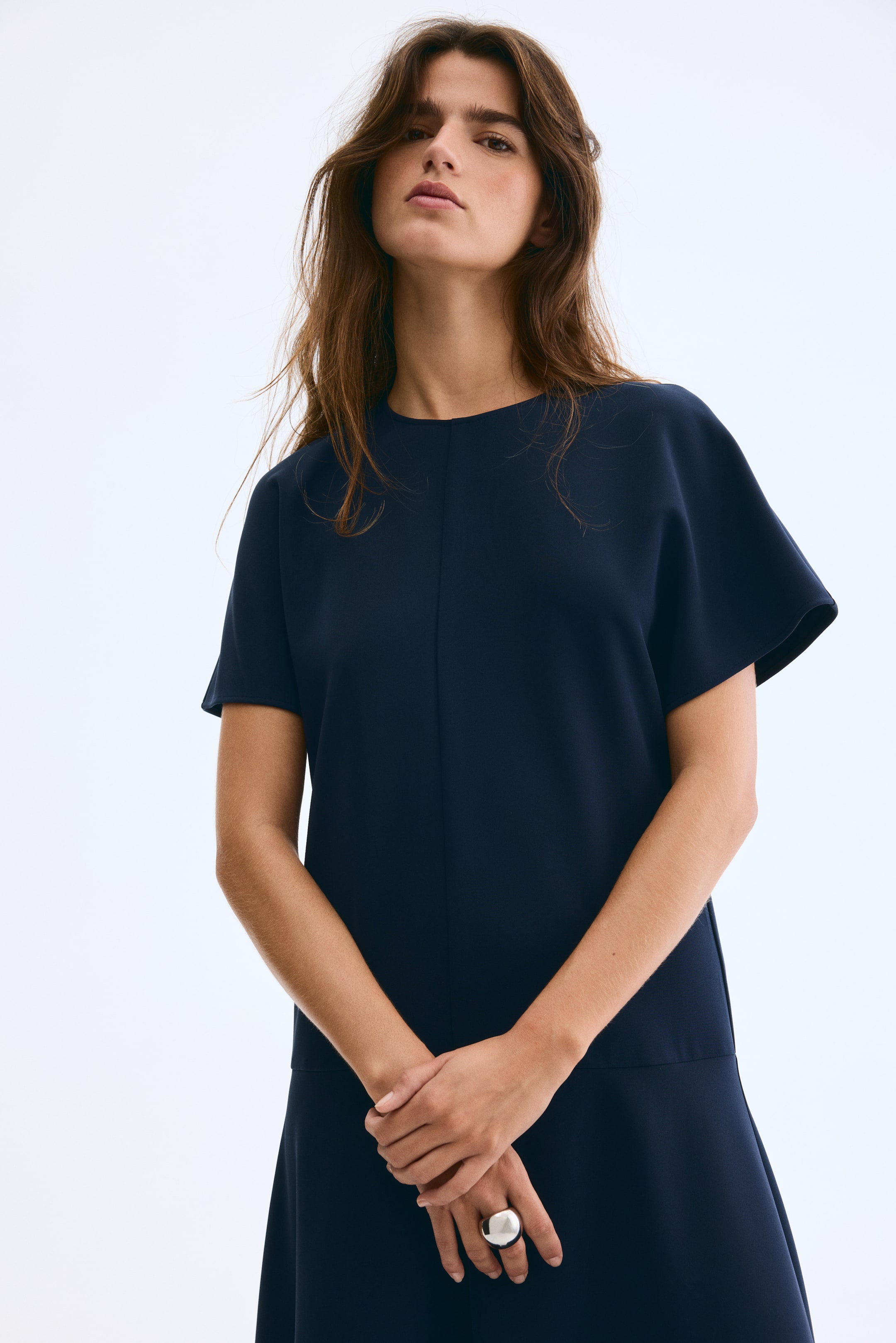 Xem ảnh lớn hơn: Một chiếc váy màu xanh hải quân được nhìn từ góc phía trước bên phải, có đường viền cổ tròn và tay áo ngắn rộng, thoáng mát. Có một đường may dọc chạy xuống phía trước, với một đường may ngang xác định điểm bắt đầu của gấu váy hơi xòe.