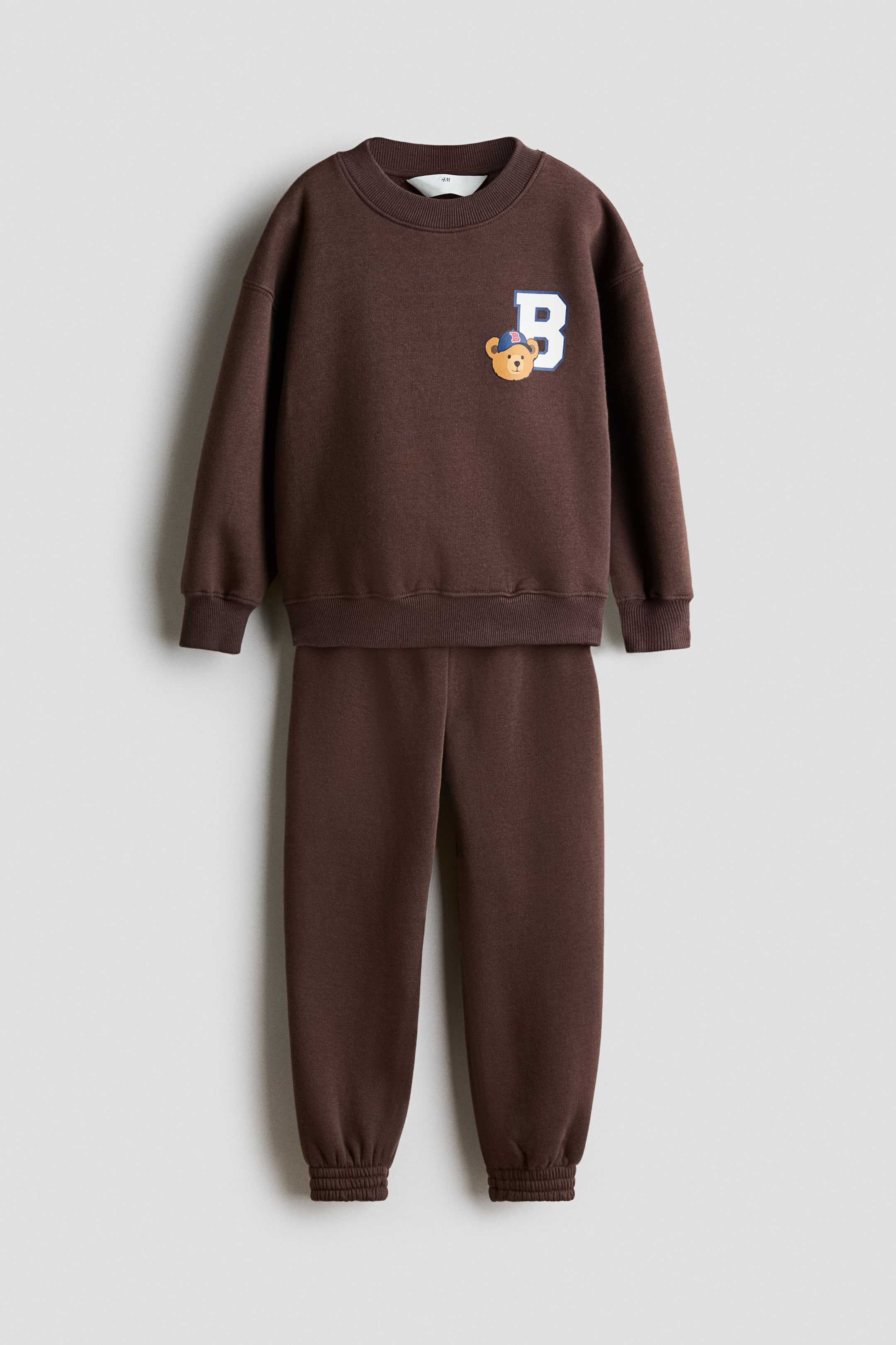 View larger image: A brown sweatshirt set, consisting of a top and joggers, is shown front-facing, with the top displaying a white 'B' letter with a blue outline and a cartoon bear head print, a round rib-trimmed neckline, dropped shoulders, and wide ribbed cuffs and hem, and the joggers featuring an elasticated waist and elasticated hems.