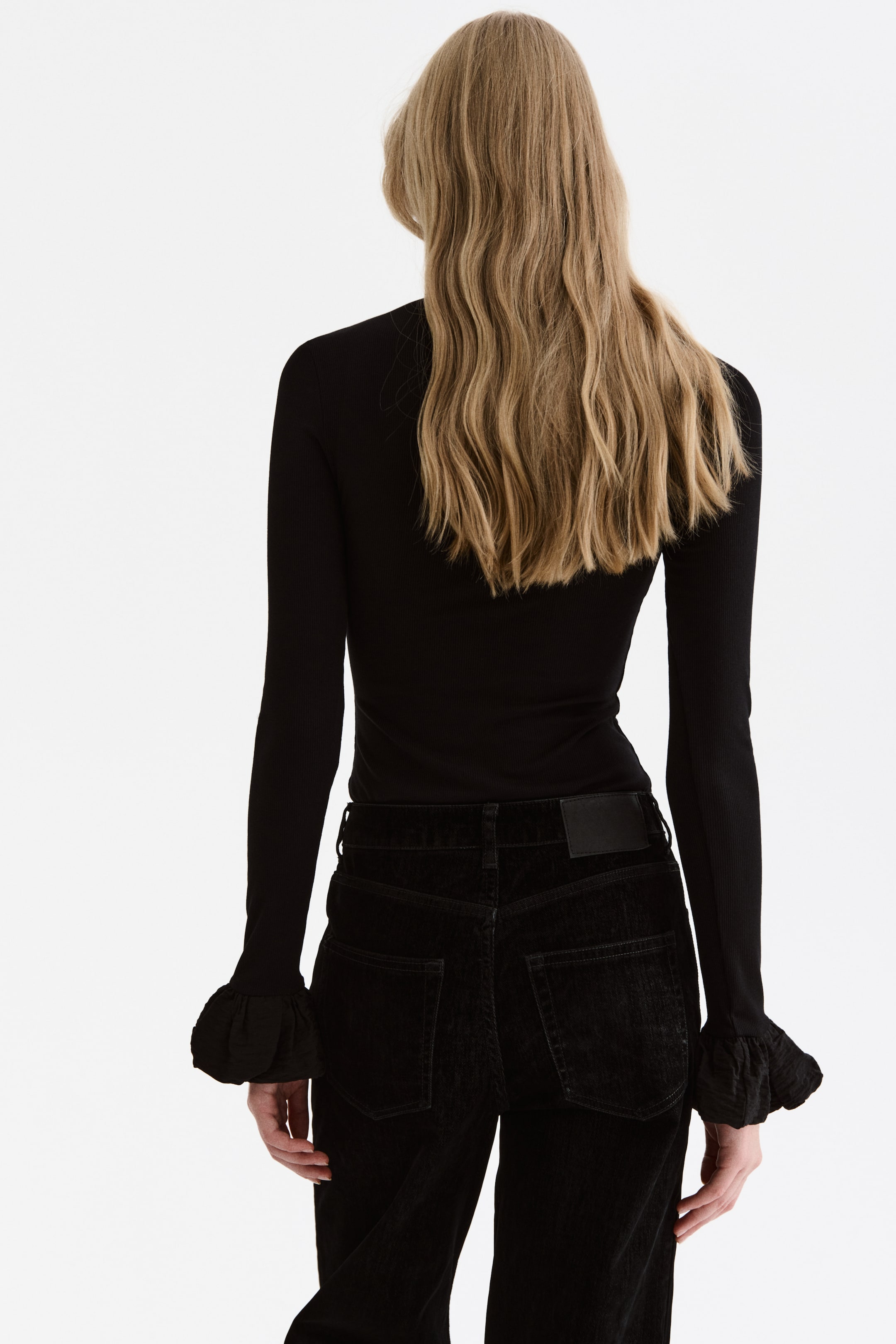 Größeres Bild anzeigen: Ein eng anliegendes schwarzes geripptes Langarmshirt mit gerüschten Manschetten wird mit einer schwarzen Cordhose getragen, die von hinten gezeigt wird. Die Hose zeigt zwei aufgesetzte Gesäßtaschen und ein schwarzes Etikett am Bund.