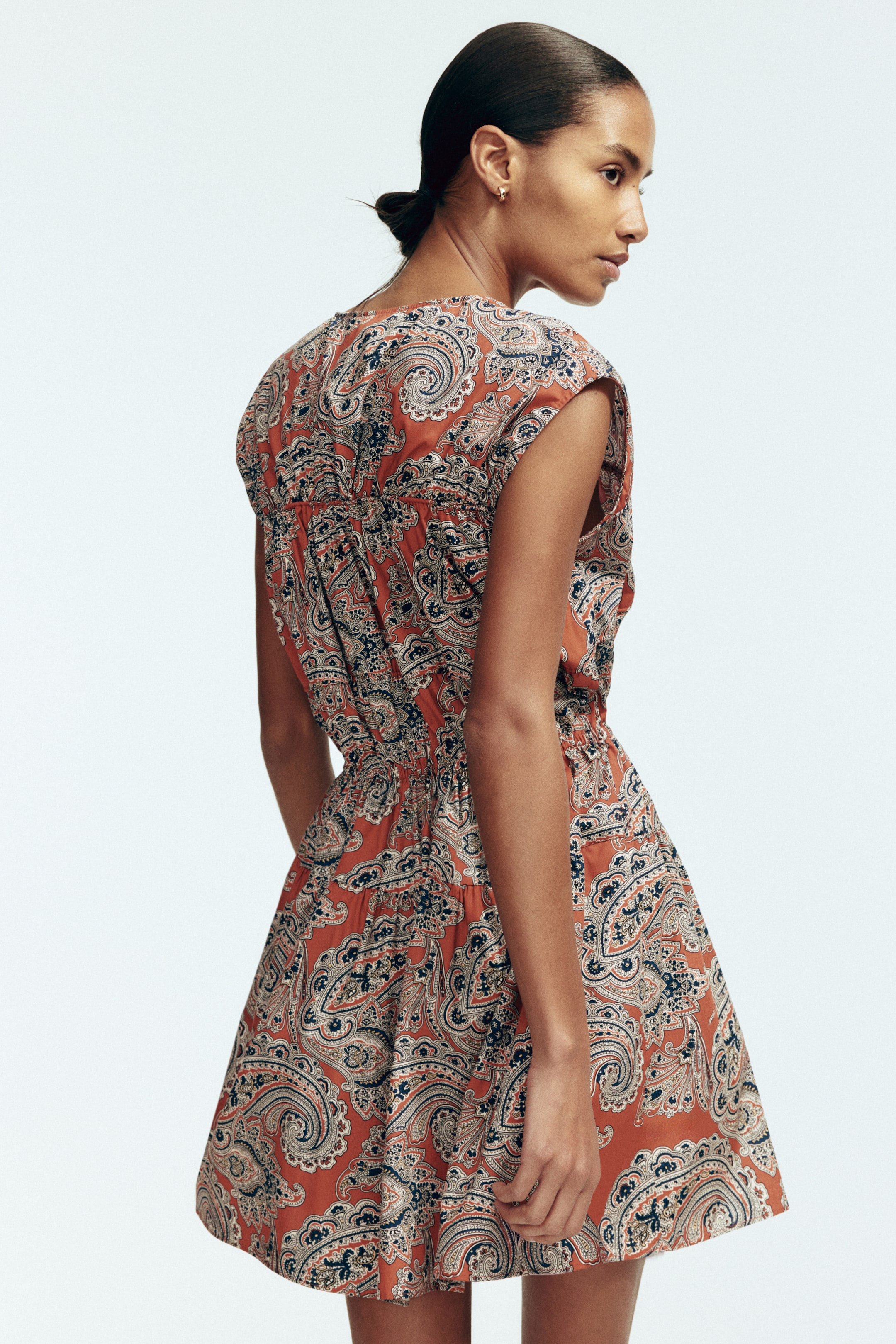 View larger image: The mini dress is featuring a terracotta orange base with a paisley print in dark blue and cream, observed from a back-right-side angle. It is designed with cap sleeves, a round neckline at the back, and a shirred waistline that is gathering the fabric into a flared skirt.