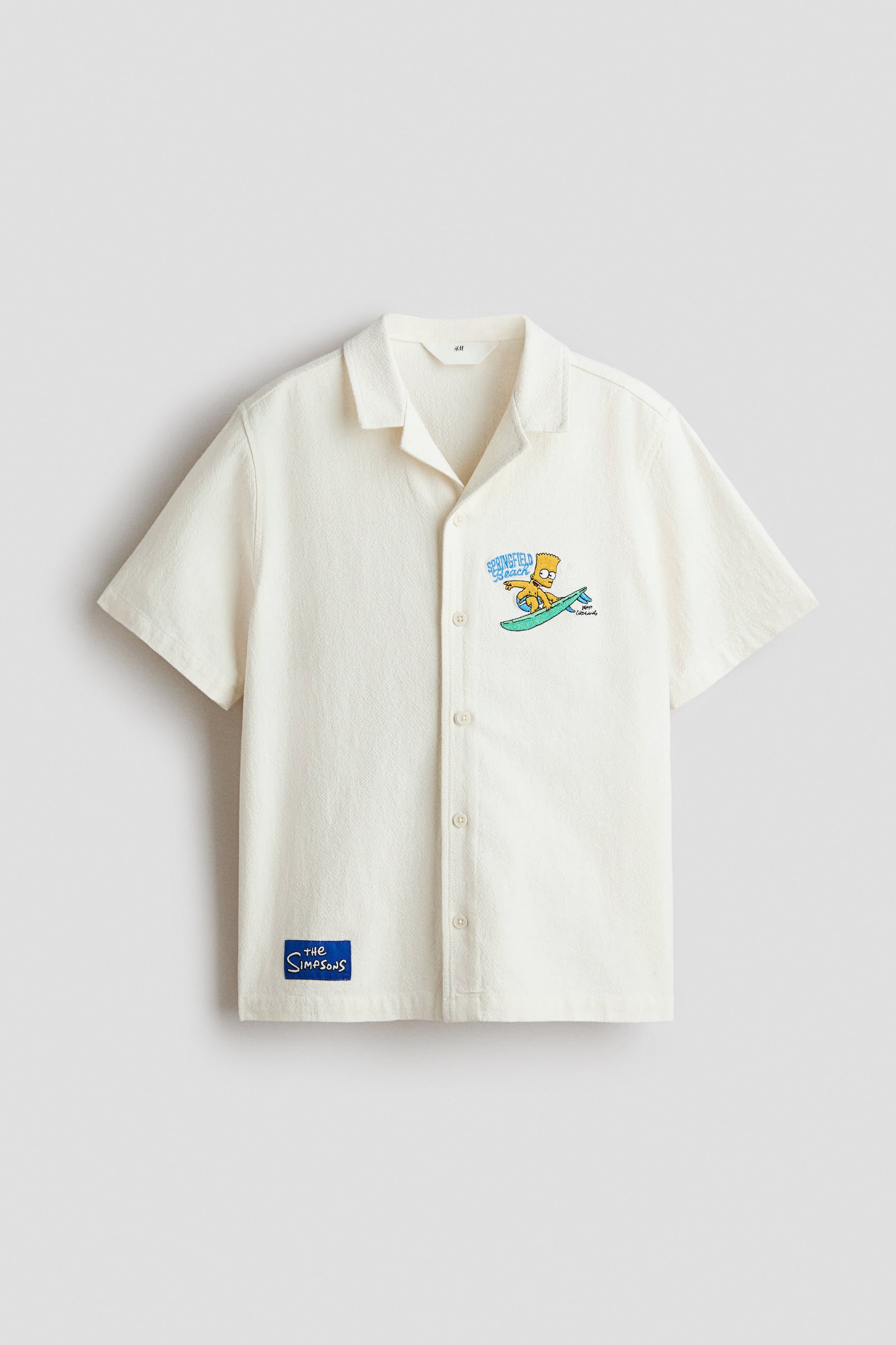 Grotere afbeelding bekijken: Een off-white overhemd met korte mouwen heeft een kampkraag en een knoopsluiting aan de voorkant. Een afbeelding van Bart Simpson die surft met de tekst 'Springfield Beach' is geborduurd op de linkerborst, en een blauw 'The Simpsons' embleem is op de linkeronderzoom genaaid.