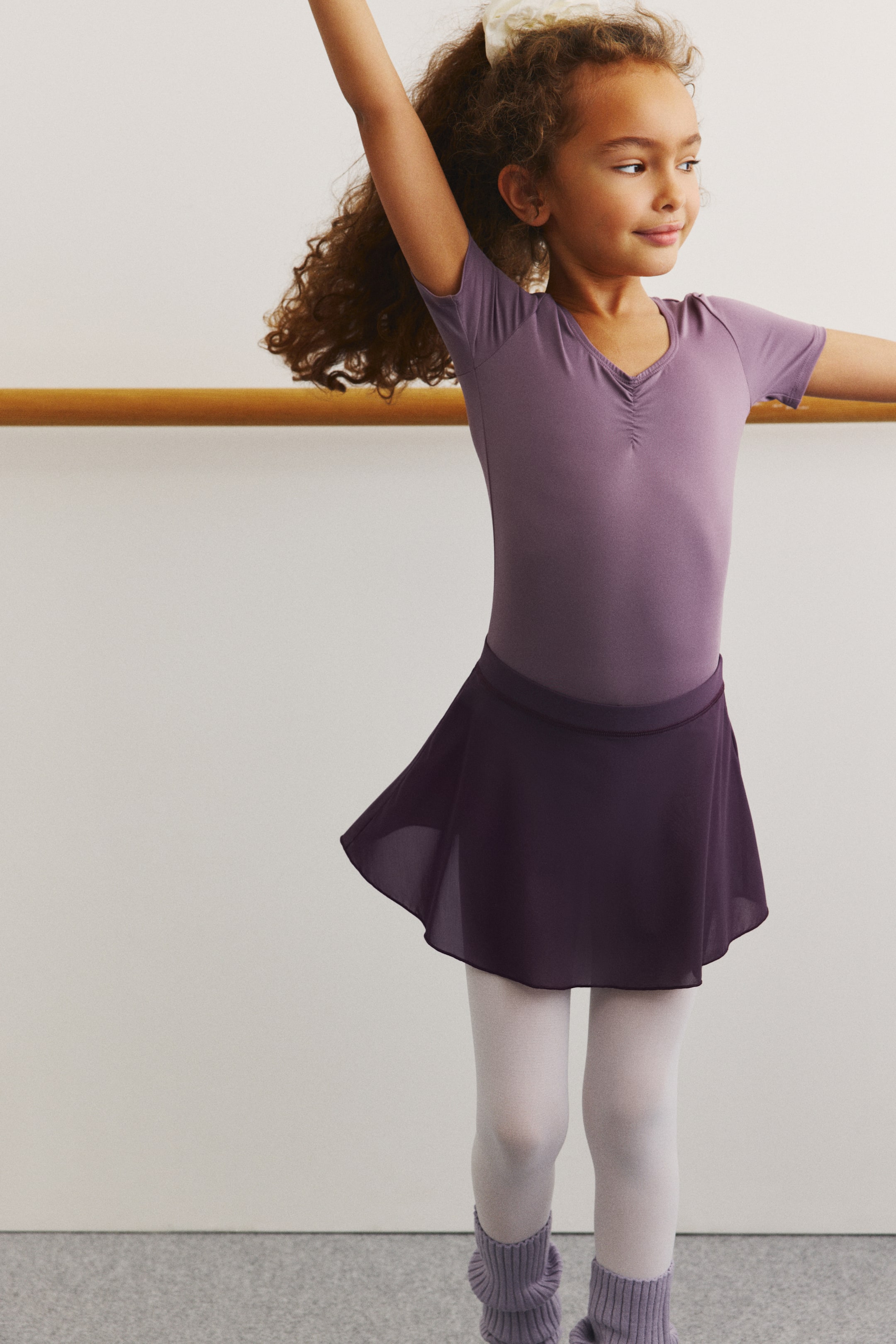 拡大画像を表示: ツートーンのバレエレオタードは、落ち着いたラベンダーの半袖トップにギャザー入りのVネック、そしてプラムパープルの身頃を特徴としています。透け感のあるプラムパープルのバレエスカートをレオタードの上に重ね、薄いラベンダーのリブ編みレッグウォーマーが白いバレエタイツを覆っています。