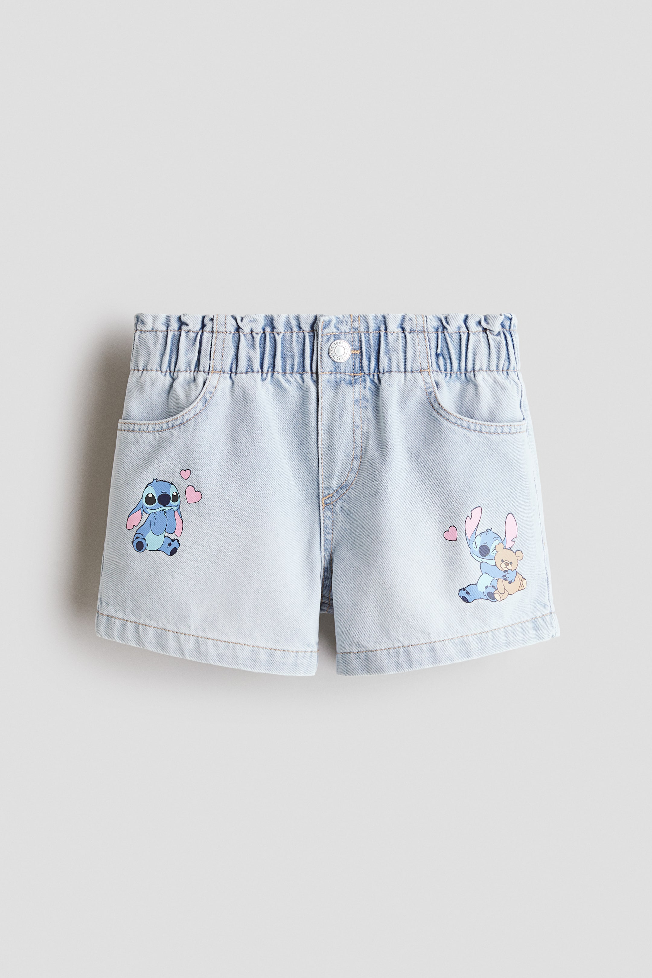 Xem ảnh lớn hơn: Quần short denim màu xanh nhạt có cạp lưng co giãn, xếp ly với một nút trắng cài và hai túi trước dạng scoop. Ống bên trái có hình vẽ Stitch với những trái tim màu hồng, và ống bên phải hiển thị Stitch đang ôm một chú gấu bông với một trái tim màu hồng.