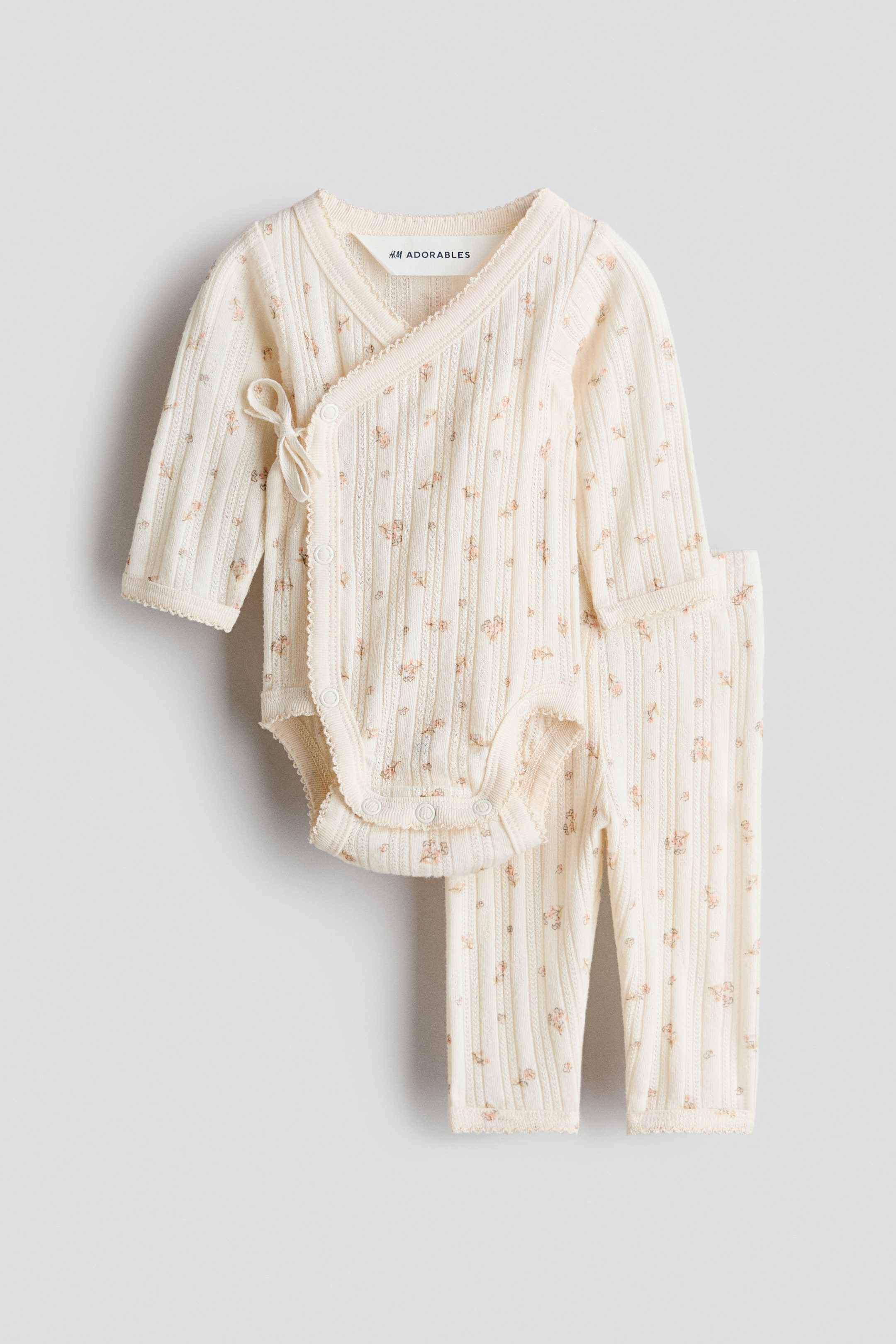 Ver imagen más grande: El conjunto H&M Adorables incluye un body de manga larga de punto jersey en color crema y leggings a juego, ambos con un pequeño estampado floral. El body, mostrado de FRENTE, está diseñado con un frente cruzado, lazos y cierres de corchete, mientras que los bordes de ambas piezas están adornados con ribetes de picot.