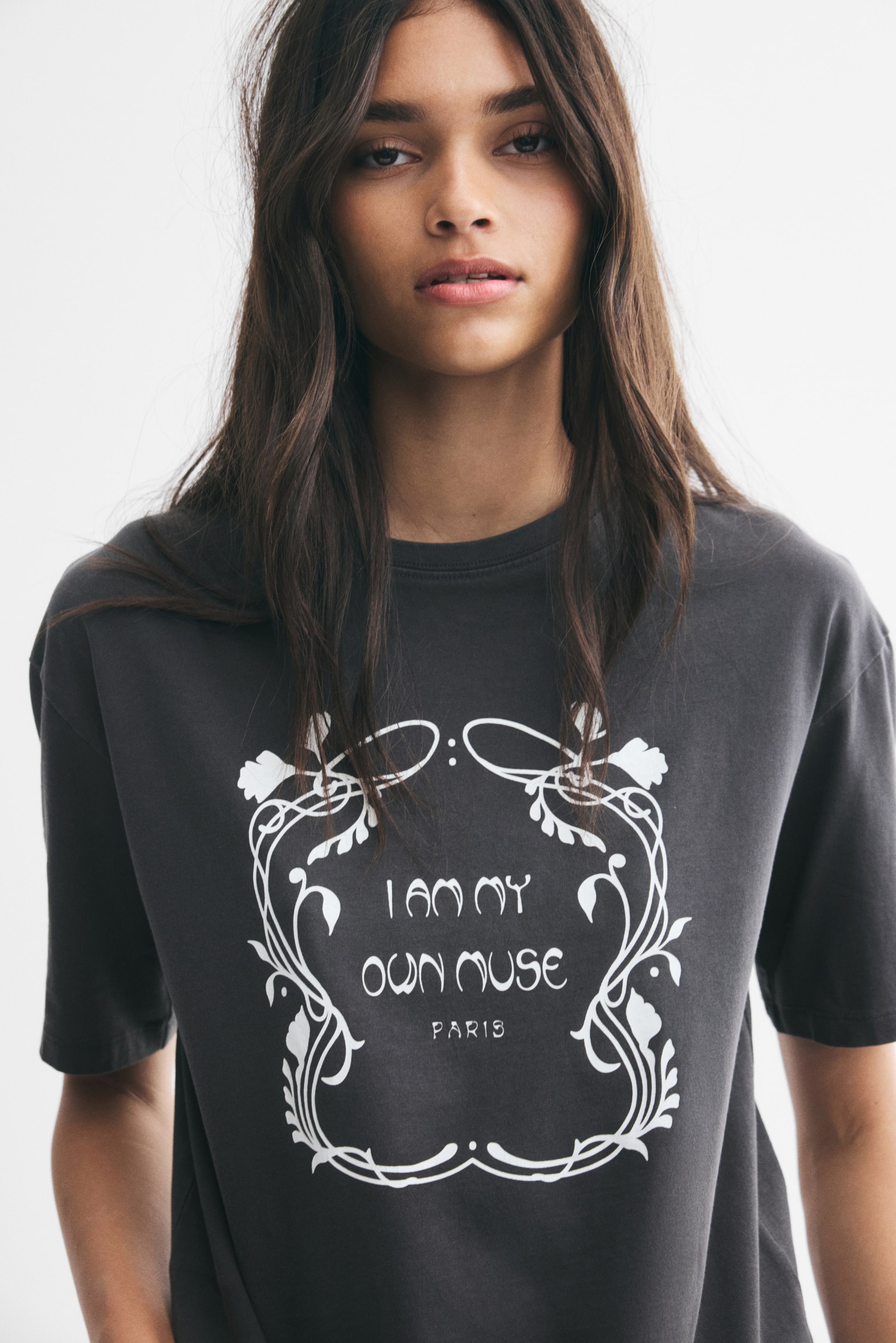 View larger image: A charcoal grey, short-sleeved T-shirt is being worn by the model, featuring a white graphic print on the front. The design displays the stylised text "I AM MY OWN MUSE" and "PARIS" enclosed within a decorative floral frame.