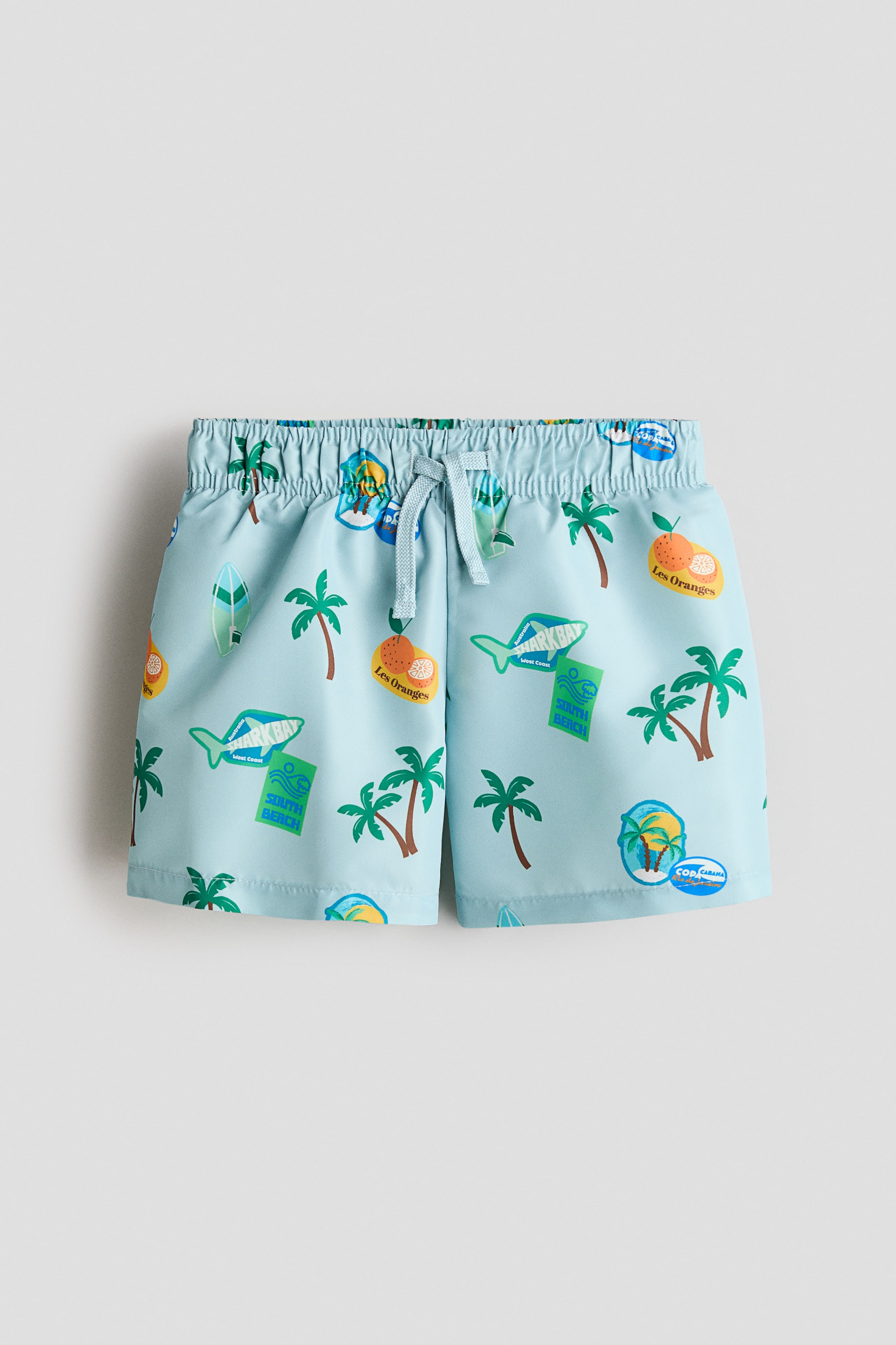 Voir une image plus grande: Shorts de bain imprimés présentés de face, arborant une couleur bleu clair avec un motif vibrant et répétitif de palmiers, de planches de surf, de requins, d'oranges et d'insignes sur le thème du voyage. Les shorts sont conçus avec une taille élastiquée et un cordon de serrage bleu clair noué à l'avant.