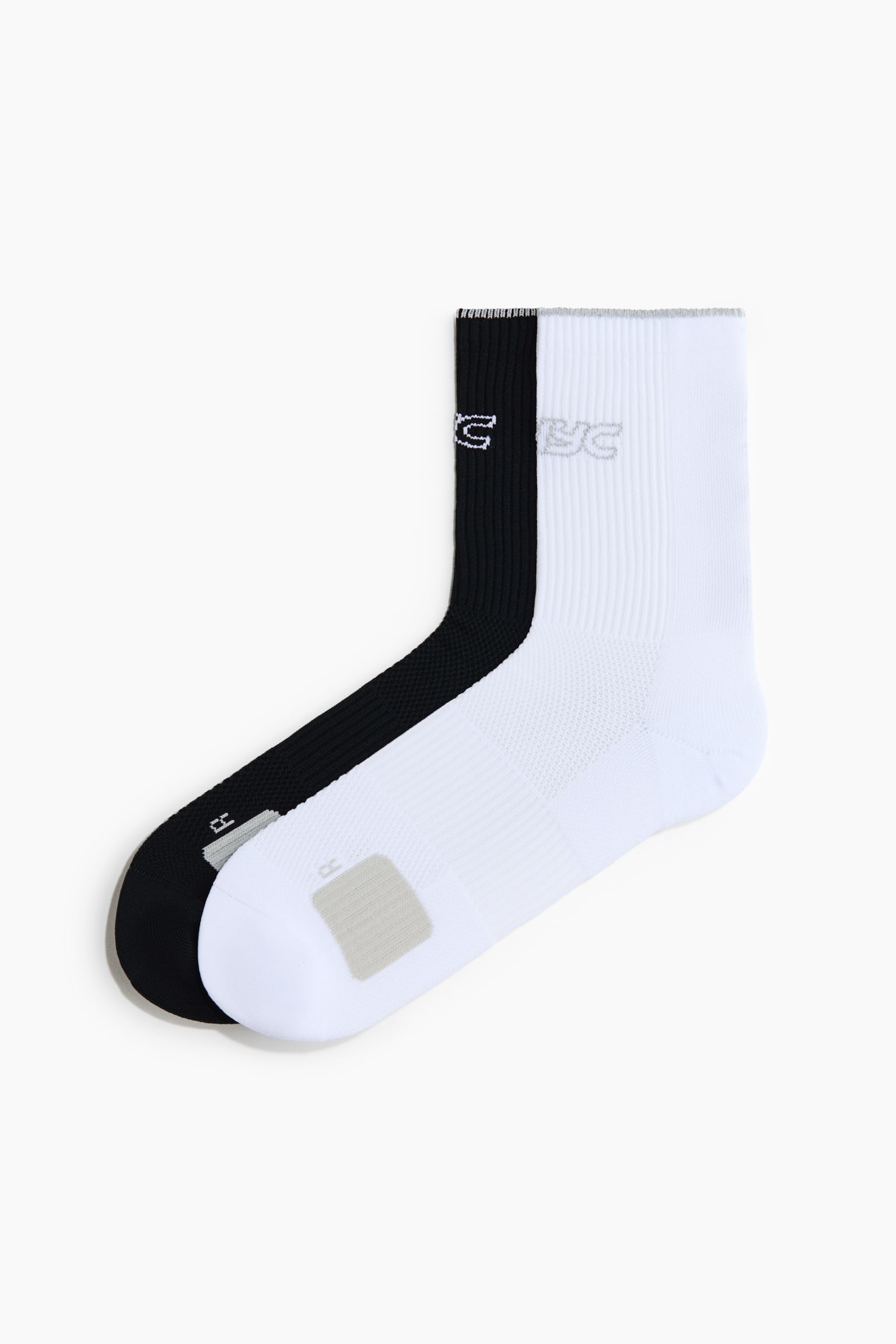 Deux paires de chaussettes de sport mi-mollet, une noire et une blanche, se chevauchent dans une vue de profil, avec des tiges cÃ´telÃ©es et une marque brodÃ©e prÃ¨s des revers. Les deux chaussettes prÃ©sentent un coussinet antidÃ©rapant rectangulaire gris et un indicateur Â« R Â» prÃ¨s des orteils.