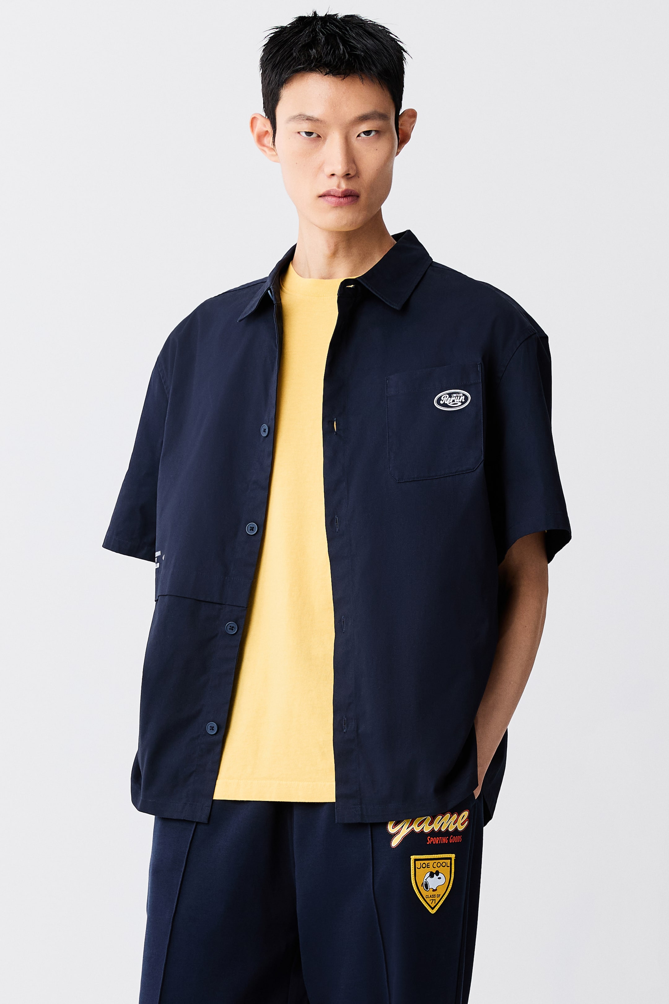View larger image: A dark blue, short-sleeve cotton shirt is worn open by the model over a yellow T-shirt. The loose-fit shirt features a classic collar, a single patch chest pocket with a small logo graphic, and a horizontal seam detail across the front.