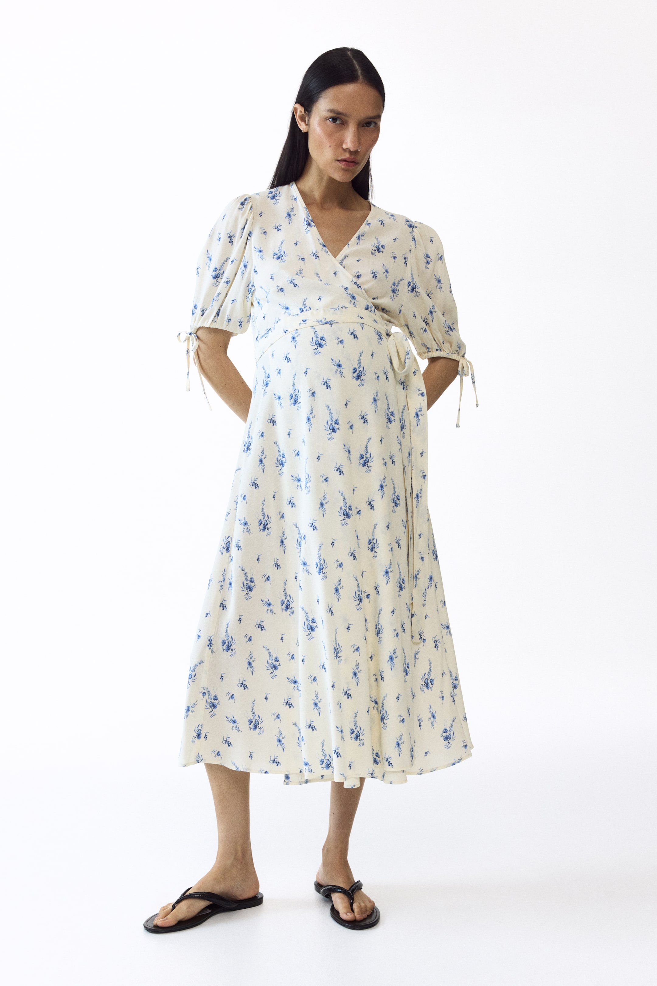 View larger image: The MAMA Puff-sleeved wrap dress is worn by the model. This cream midi dress features a blue floral print, a V-neckline, puff sleeves tied at the cuffs, and a side-tie wrap closure that cinches the waist.