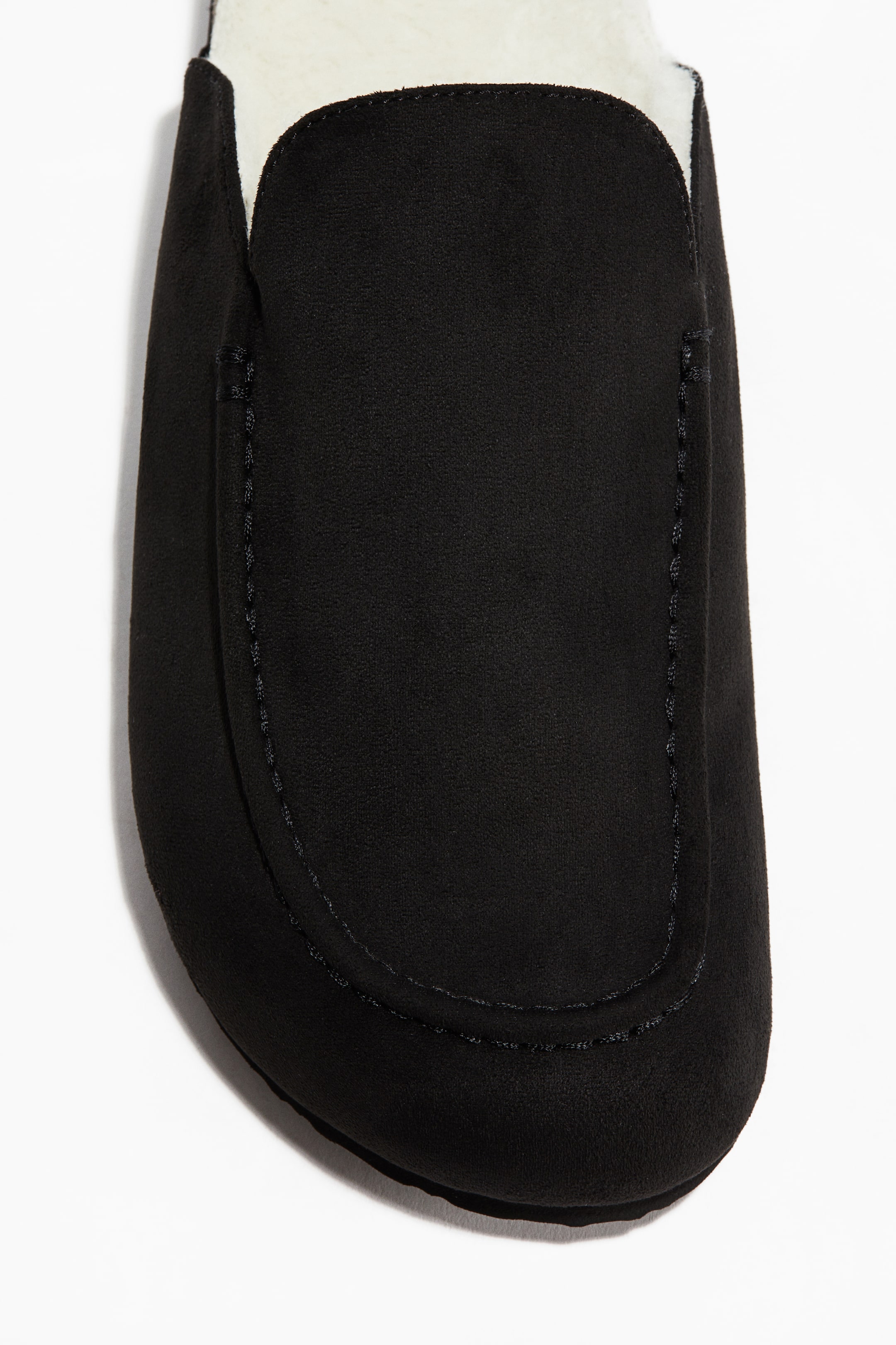 View larger image: A black moccasin-style slipper is presented from the front, showing a U-shaped topstitch across its vamp and a white plush lining along the opening.