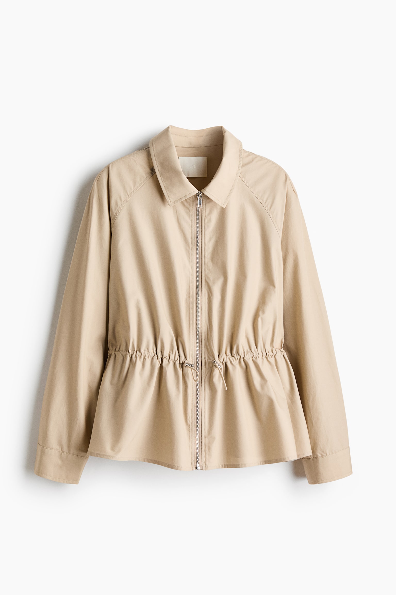 A beige cotton-weave jacket is hanging facing the front, featuring a classic collar, a silver front zipper, and long raglan sleeves. An adjustable drawstring with cord stoppers is cinching the waist, creating a flared peplum hem.