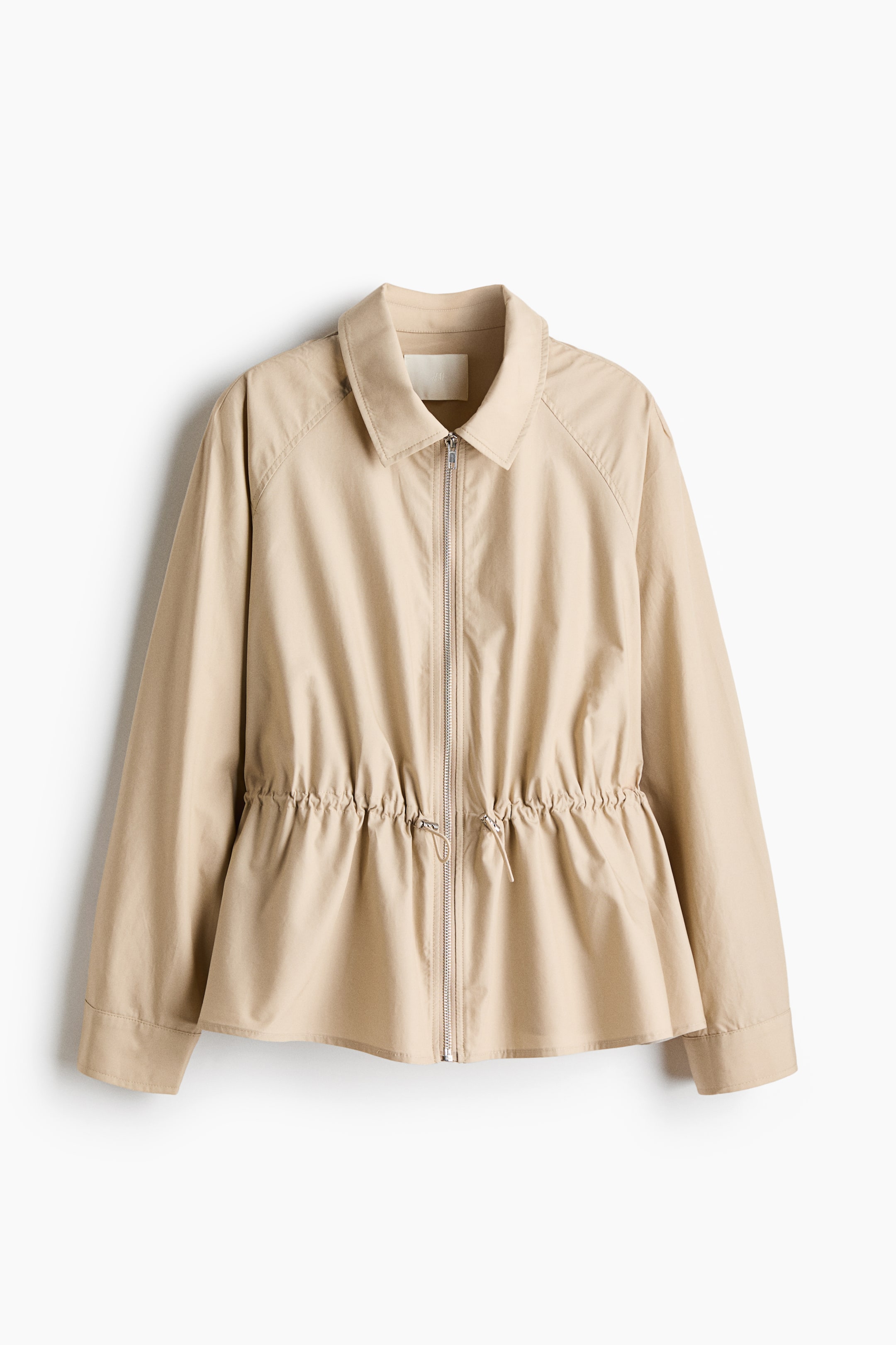 View larger image: A beige cotton-weave jacket is hanging front-facing, featuring a classic collar, a silver front zip, and long raglan sleeves. An adjustable drawstring with cord stoppers cinches the waist, creating a flared peplum hem.