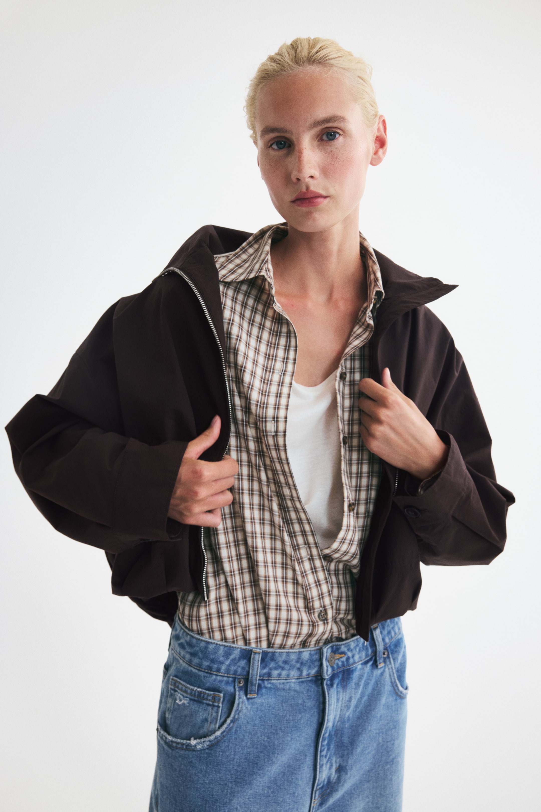 Agrandir l'image: Une veste courte oversize en tissu tissé marron foncé, avec un col montant, des épaules tombantes, des manches longues et un ourlet ballon élastiqué, est portée par le mannequin. La fermeture éclair sur le devant de la veste est partiellement ouverte, et le mannequin tient les pans avant, laissant apparaître une chemise à carreaux et un haut blanc en dessous.