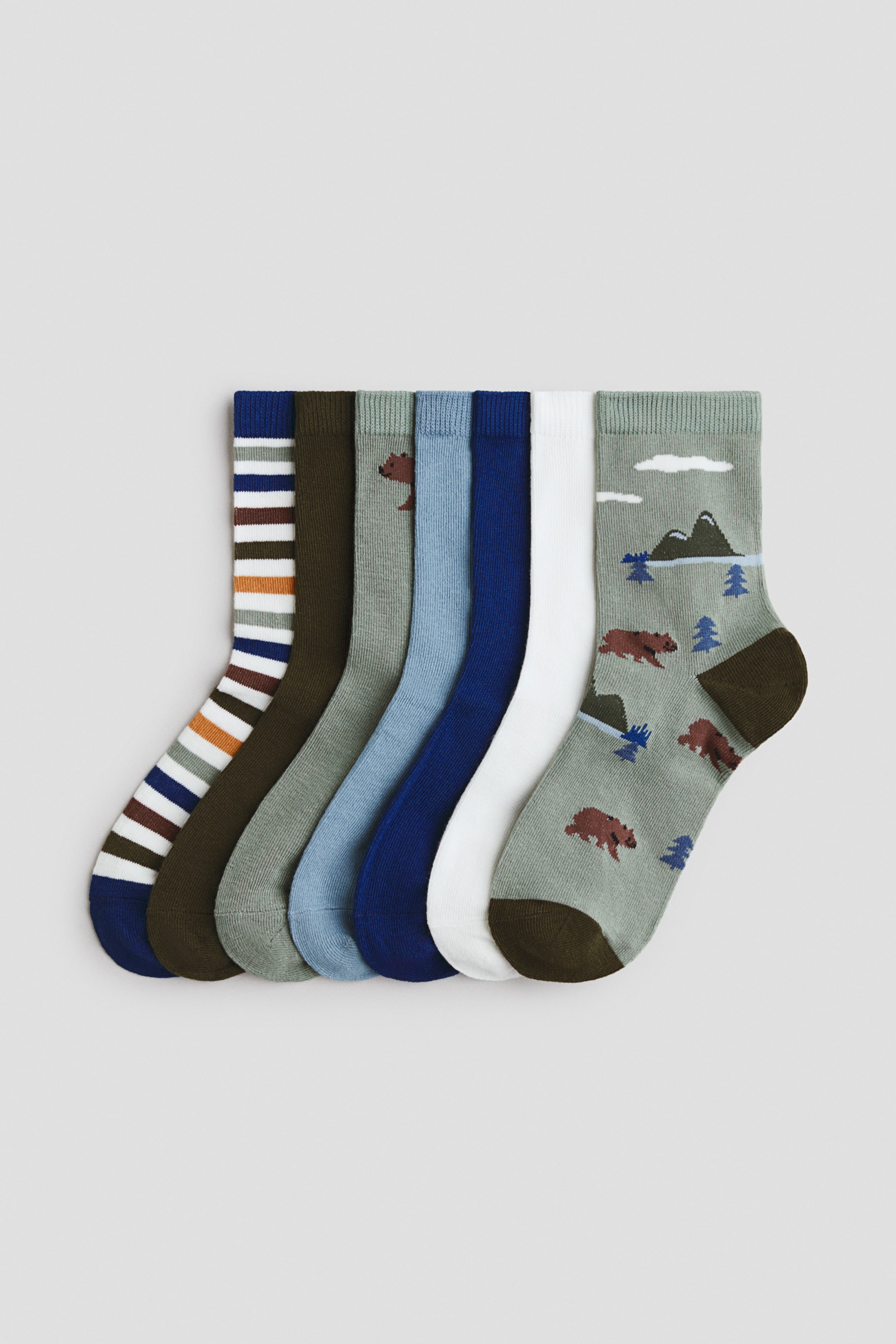 View larger image: A set of seven crew socks is displayed, fanned out from left to right. The collection includes one sock with white, brown, and blue stripes, and five solid-coloured socks in dark olive green, light olive green, light blue, dark blue, and white. A seventh light sage green sock features a pattern of brown bears, green mountains, and blue trees, with dark olive green toes and heels.