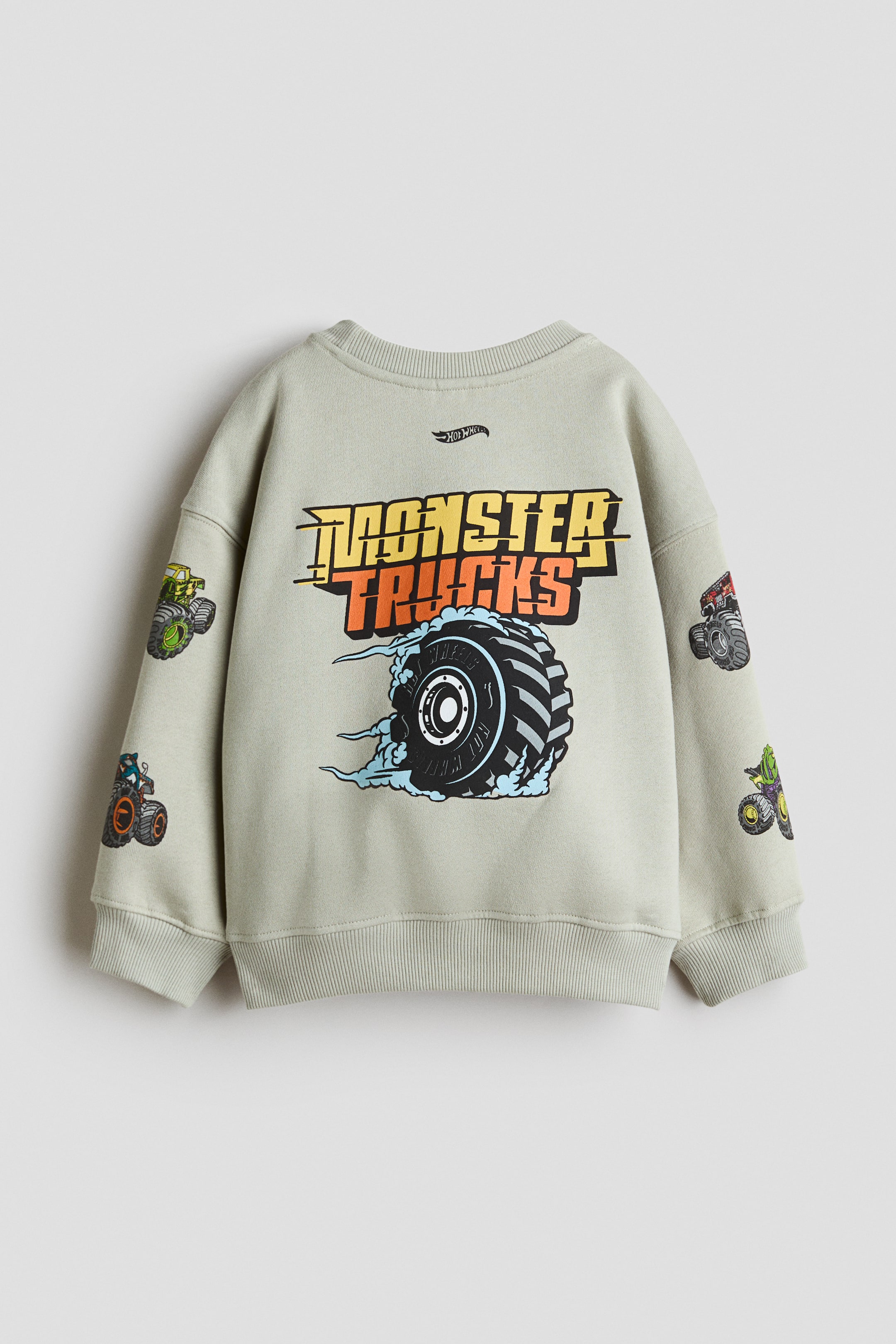 Se større billede: En lysegrå sweatshirt med rund hals viser bagsiden og har en stor grafik med teksten "MONSTER TRUCKS" i gult og orange over en illustration af et snurrende dæk. Sweatshirten har sænkede skuldre, ribkanter ved ærmerne, en ribkant forneden og mindre monstertruck-grafikker trykt langs begge ærmer.