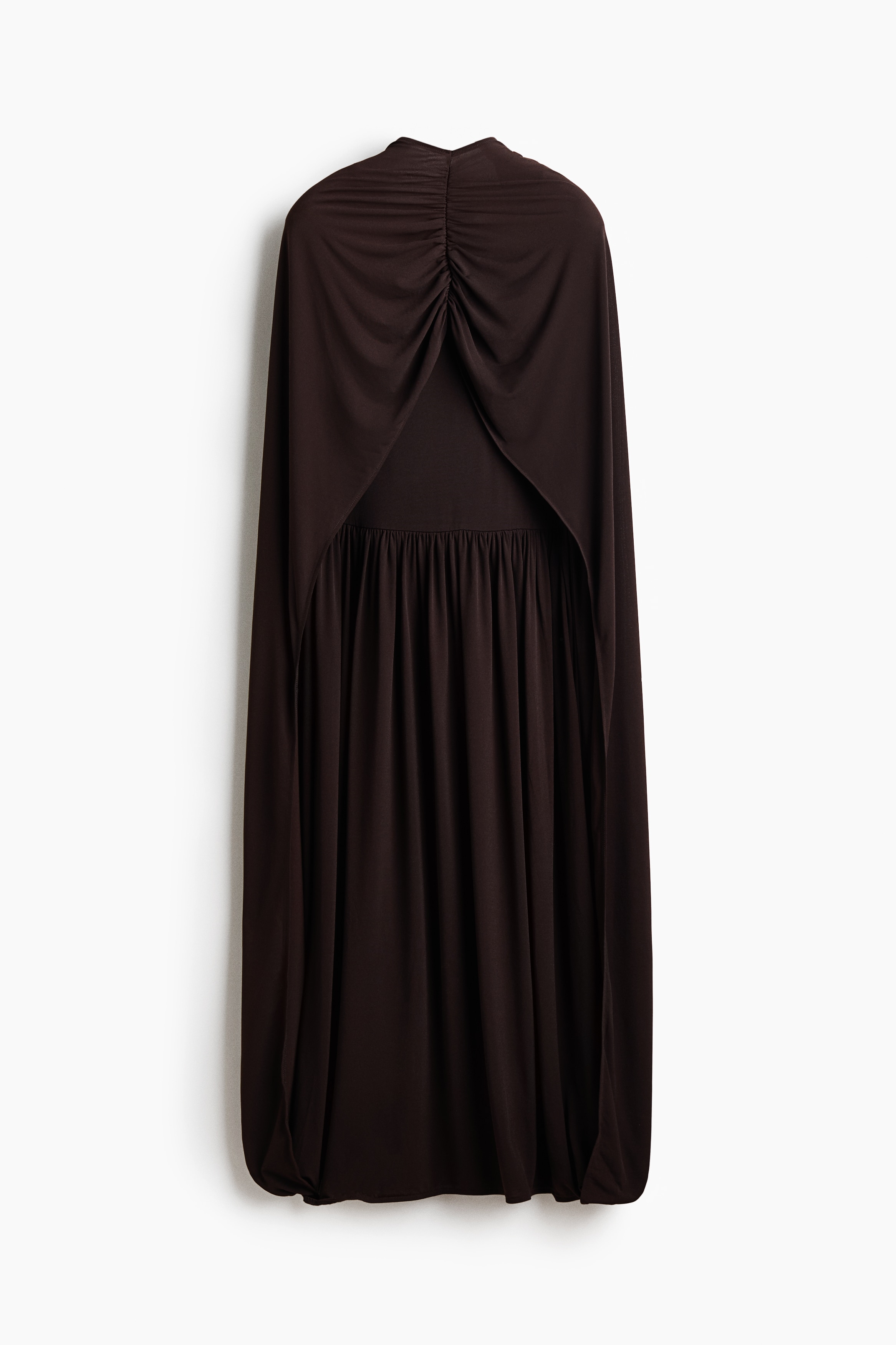 The model is wearing a dark brown jersey dress featuring a cape-like outer layer with a gathered neckline that drapes over the shoulders and arms. The inner dress layer has a gathered waist and is styled with black heeled sandals.
