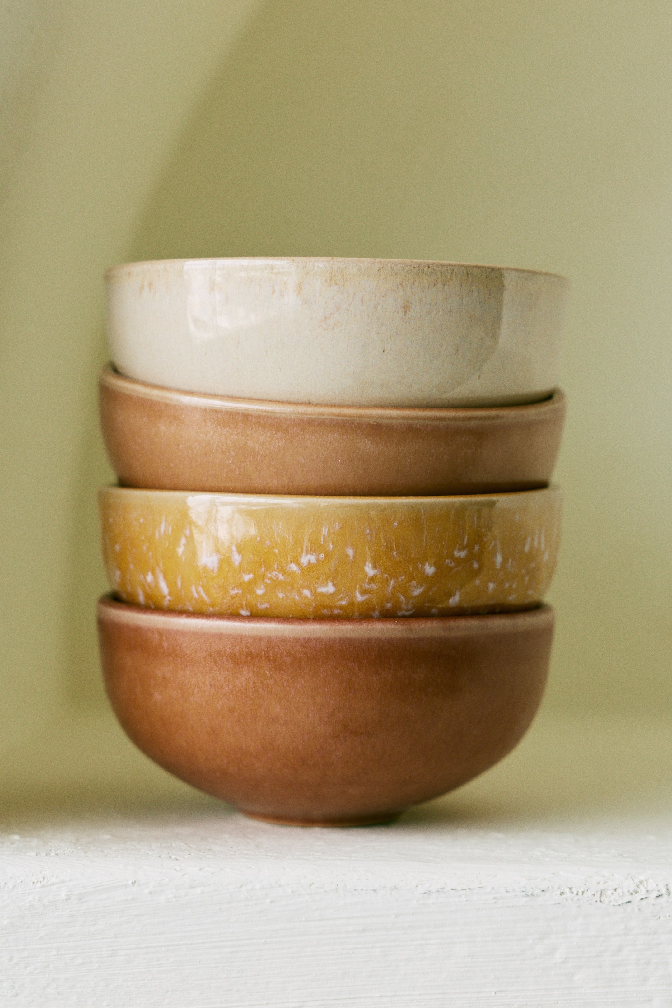 Visualizza immagine più grande: Quattro ciotole rotonde in ceramica sono impilate verticalmente, ognuna con una smaltatura distinta. Dall'alto verso il basso, le ciotole sono presentate in beige chiaro, marrone medio solido, giallo maculato e marrone scuro solido.