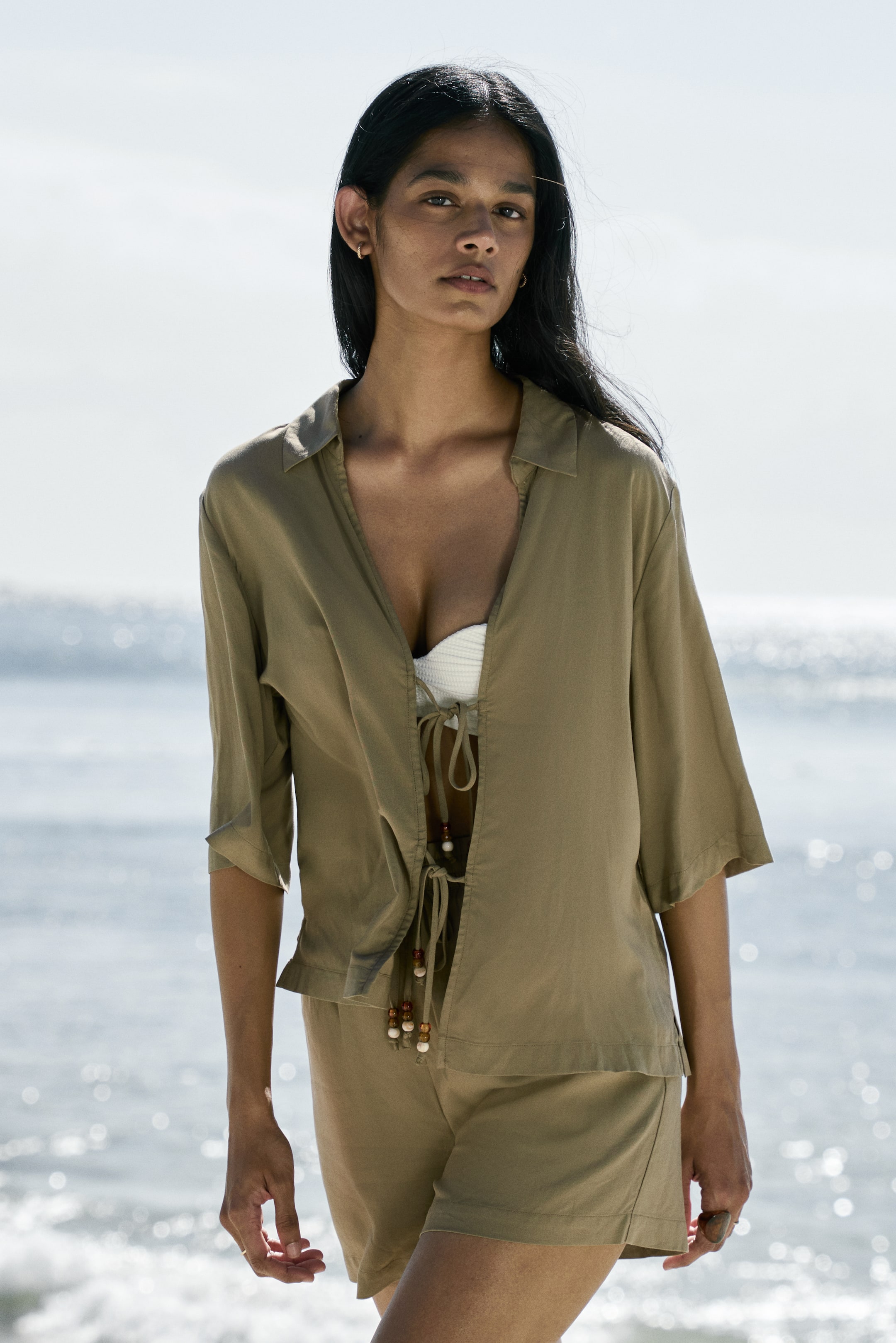 View larger image: A front-facing khaki loose-fitting shirt is worn open over a white ribbed bralette, featuring a pointed collar, elbow-length sleeves, and front ties with decorative wooden beads. Matching khaki high-waisted shorts accompany the look, completing this coordinated set.
