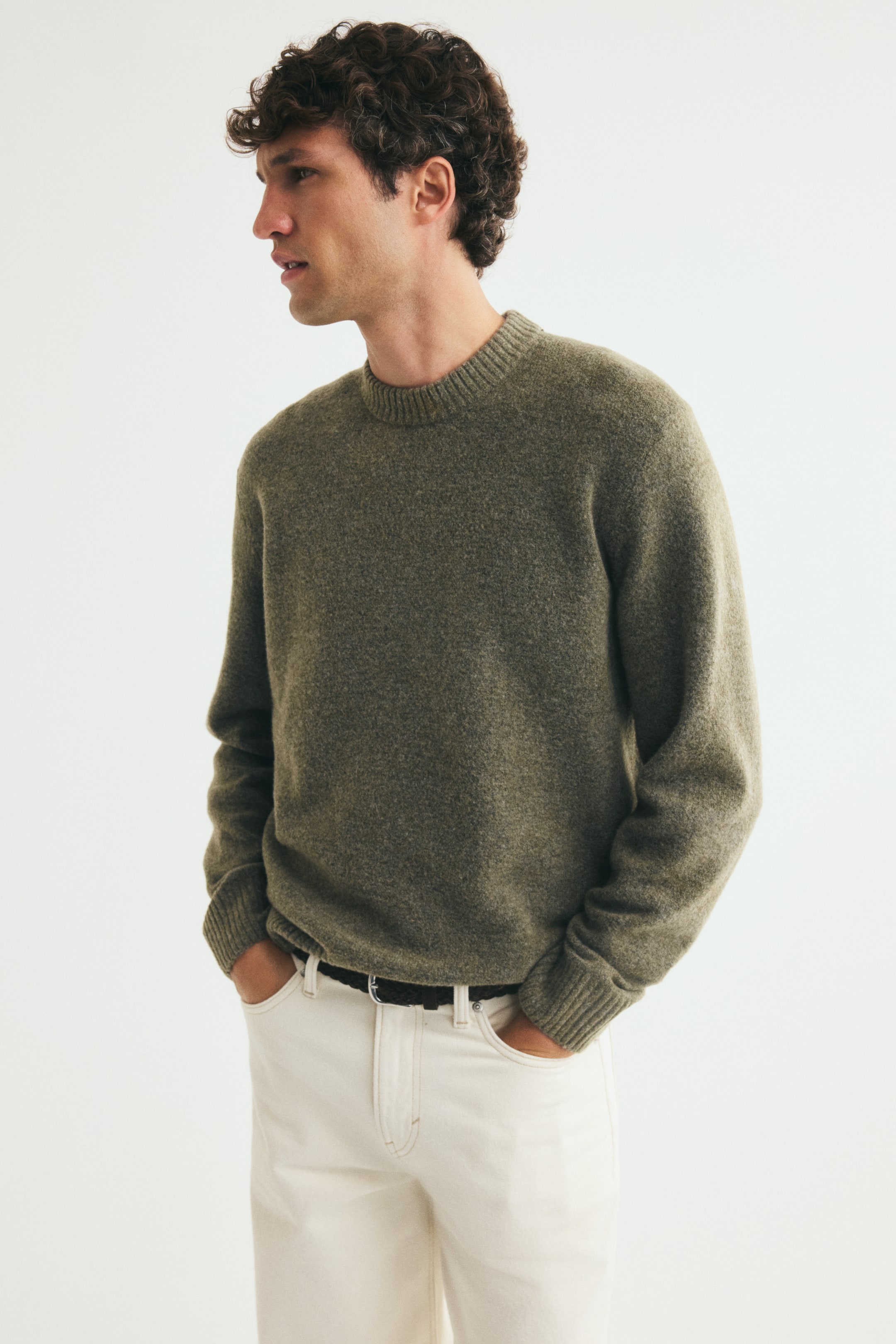 View larger image: A green, brushed-knit sweater featuring a ribbed crew neckline, ribbed cuffs, and a ribbed hem is being worn by the model. The sweater is styled tucked slightly into off-white trousers with a dark belt, with the model resting their hands in the pockets.