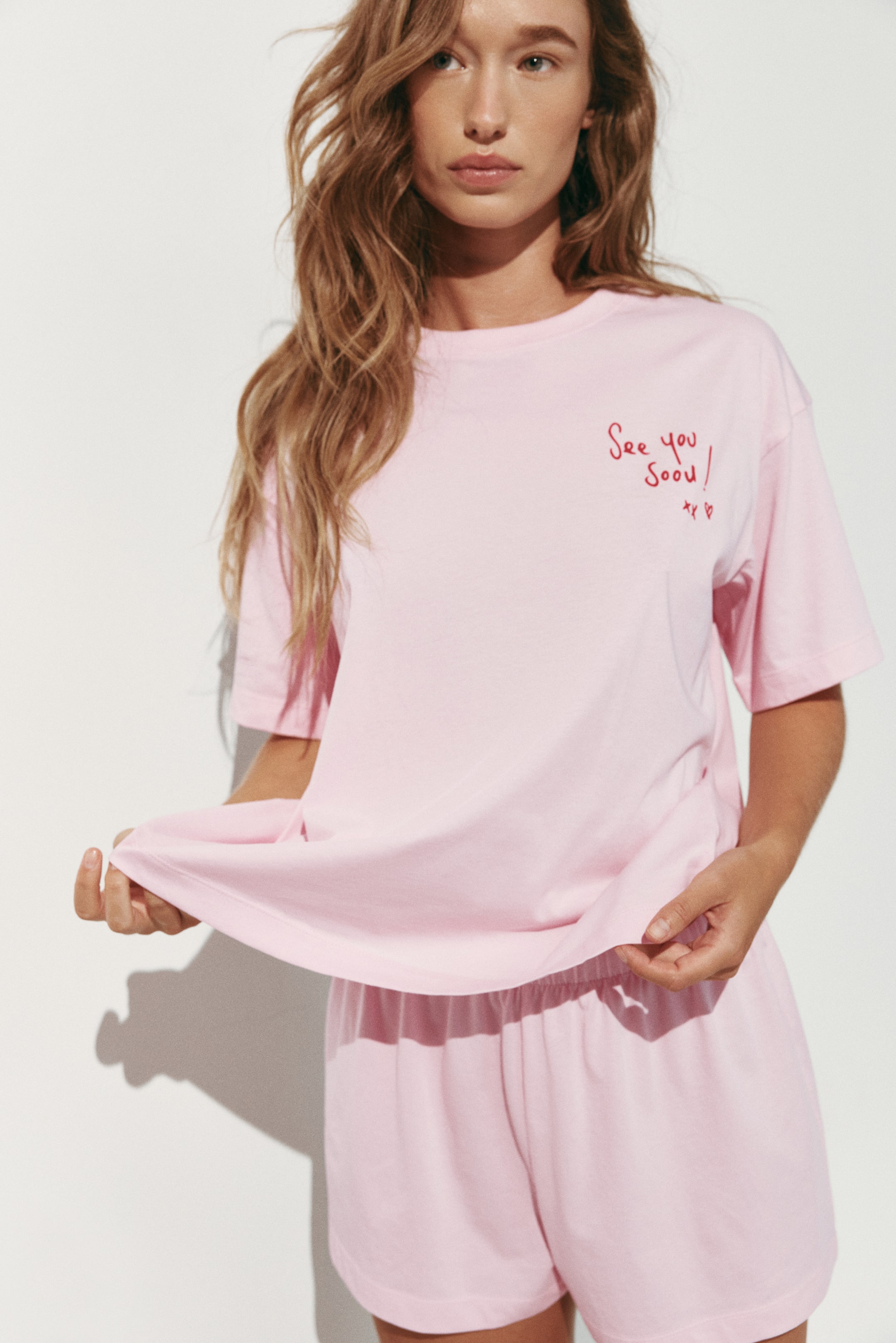 Visualizza immagine più grande: Un pigiama in jersey di cotone rosa chiaro è indossato frontalmente, composto da una T-shirt dalla vestibilità morbida con spalle scese, girocollo a costine e scritta rossa stampata sul lato sinistro del petto. I pantaloncini coordinati presentano una vita elasticizzata coperta.