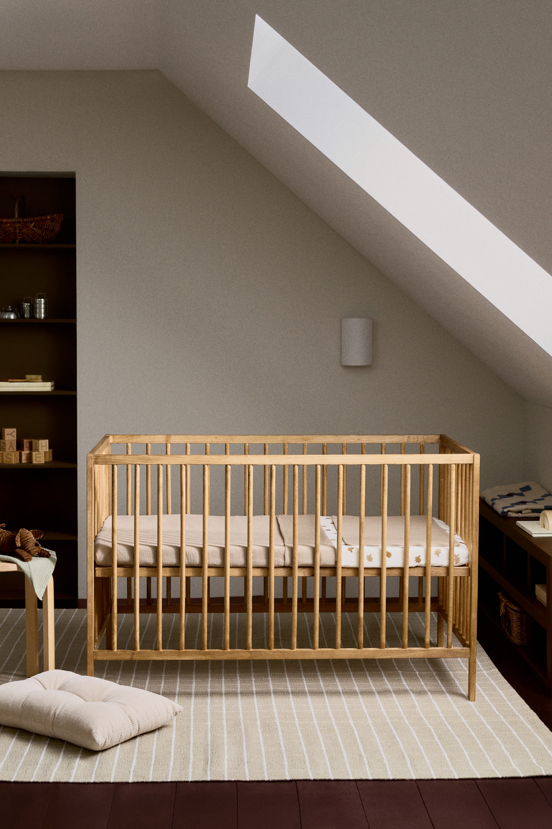 Nagyobb kép megtekintése: Egy világos tónusú fa kiságy enyhe szögben van bemutatva, látható az eleje és a jobb oldala. Függőleges lécekkel rendelkezik, és világos bézs színű, finom mintázatú lepedővel van felszerelve, amelyen egy sima bézs takaró van áthajtva.