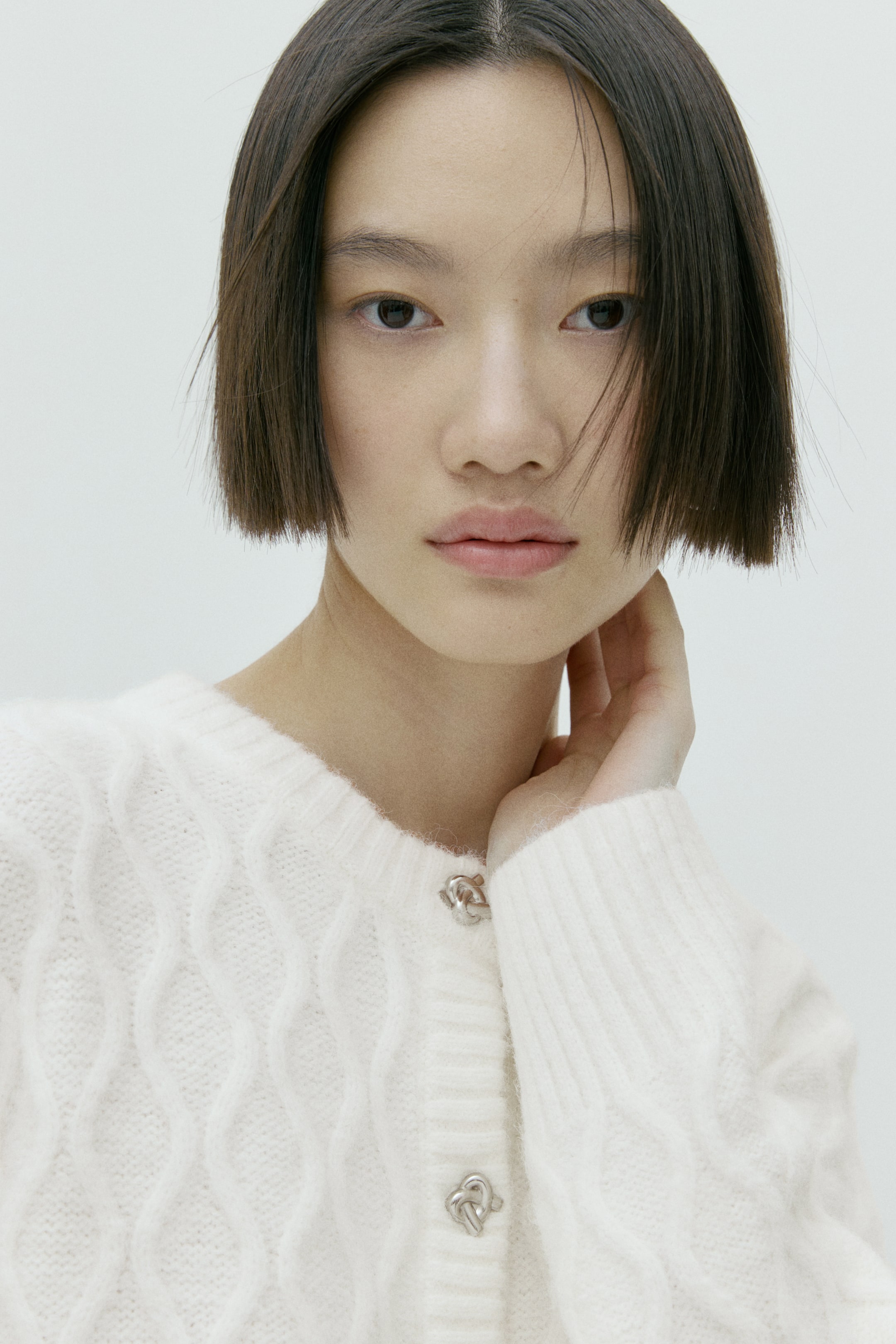 拡大画像を表示: 白のカーディガンは胴部分に凹凸のあるダイヤ型のケーブル編みを施し、プレーンニットの袖とコントラストを成しています。装飾的なシルバー調の絡み合う結び目モチーフのボタンが前立てを留め、手が首元にそっと触れてネックラインを際立たせています。