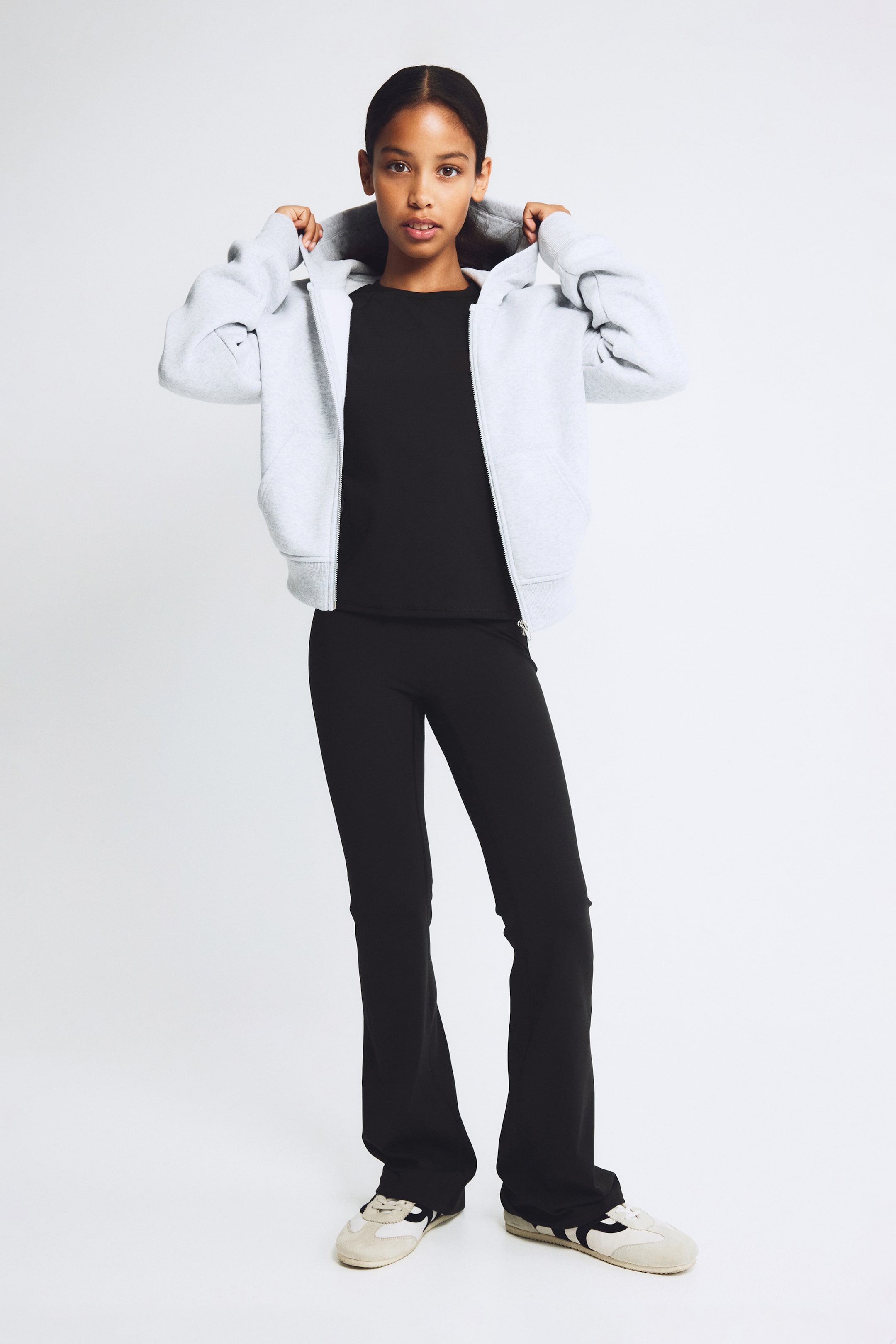 Grotere afbeelding bekijken: Een lichtgrijze hoodie met rits, afgebeeld van voren, wordt open gedragen over een zwart crewneck T-shirt en een zwarte flared broek. De hoodie heeft een ruime capuchon, een zilverkleurige ritssluiting aan de voorkant en ribboorden aan de mouwen en onderzoom.
