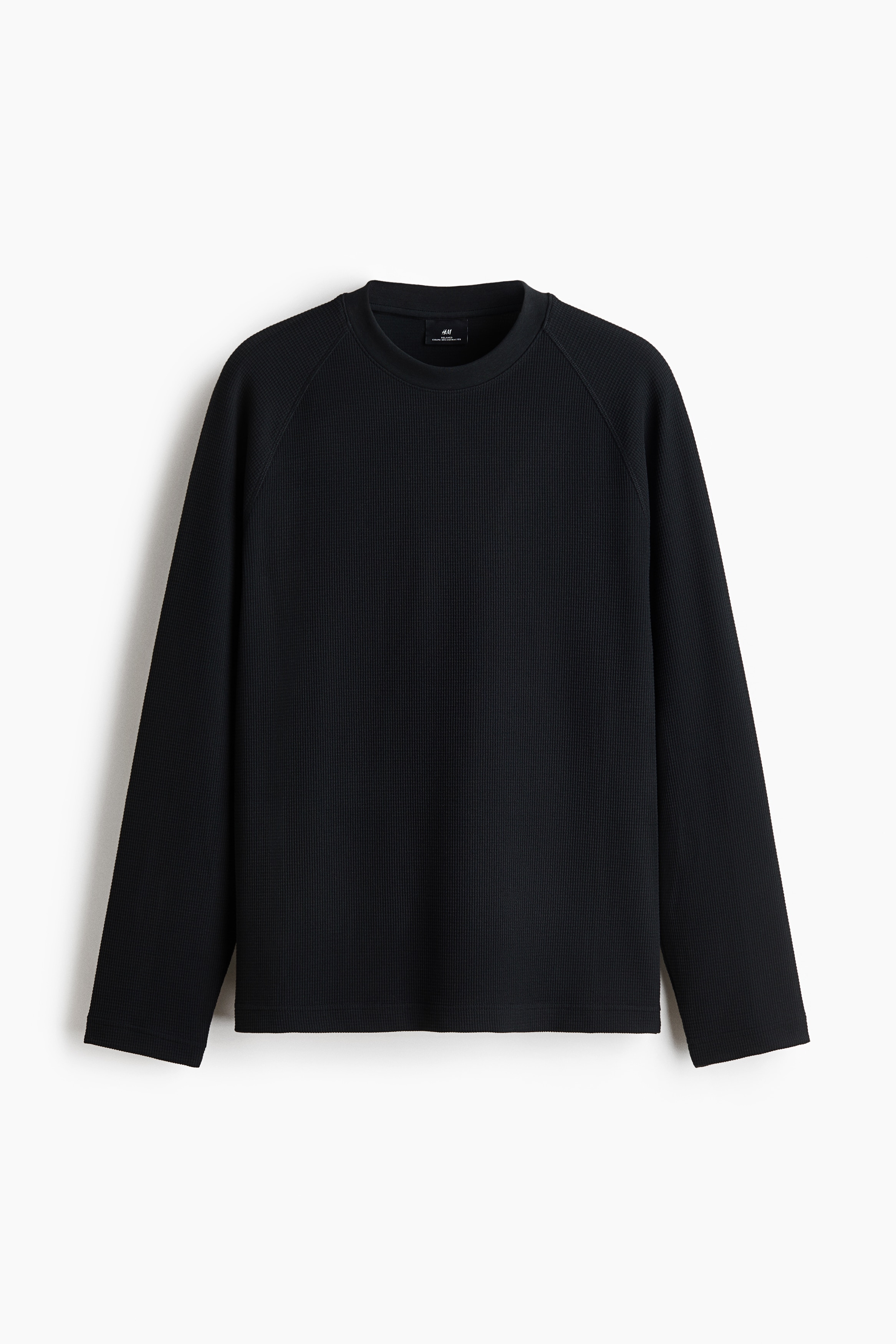 Un t-shirt en jersey noir texturÃ© Ã  motif gaufrÃ©, Ã  coupe dÃ©contractÃ©e, avec encolure ronde et manches raglan longues, portÃ© par le mannequin. Le t-shirt est agencÃ© avec un pantalon noir assorti Ã  cordon de serrage.