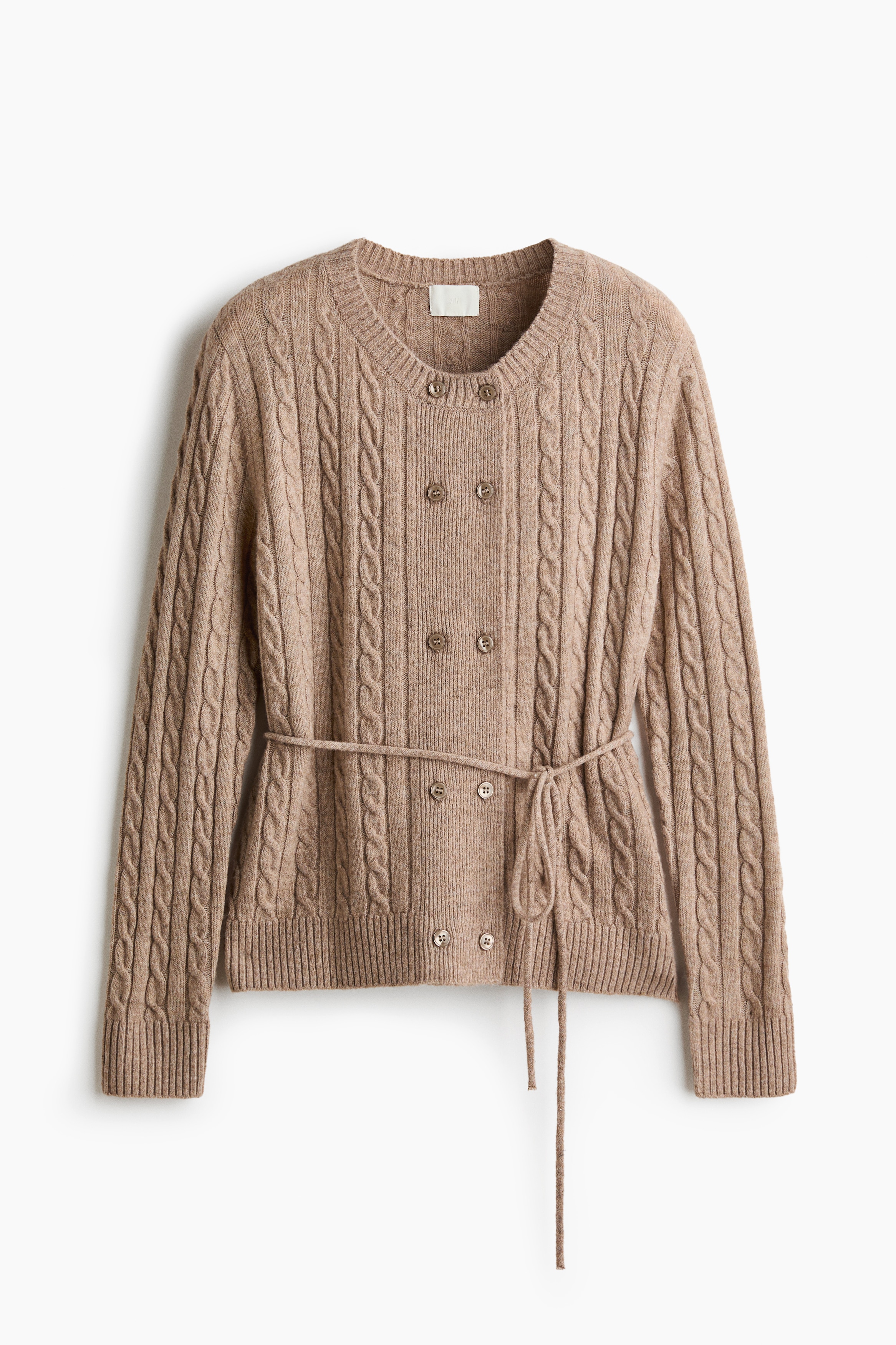 A beige cable-knit cardigan with a round ribbed neckline, double-breasted button closures, and a narrow waist tie belt is worn by the model. The cardigan is layered over a white shirt with a scalloped collar and paired with dark trousers.