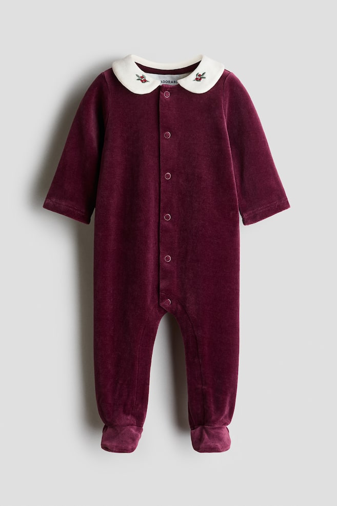 A burgundy velour baby sleepsuit is shown from the front, featuring a white Peter Pan collar detailed with small embroidered holly motifs. The long-sleeved, footed garment features a central row of press studs running down the front. - 1