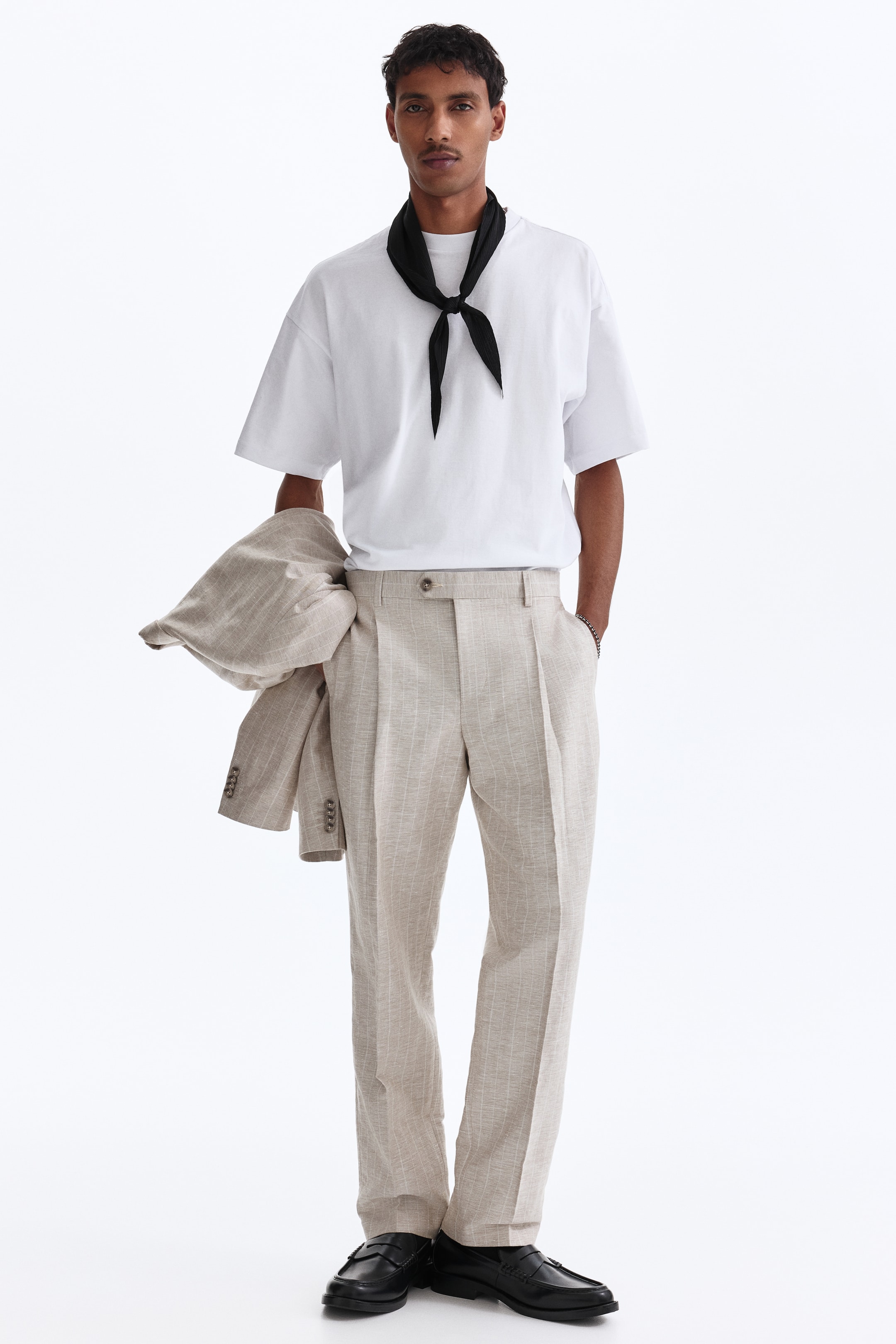 View larger image: An oversized white T-shirt is worn with light beige pinstripe pleated trousers. A black neckerchief is tied at the model's neck, and a matching pinstripe jacket is carried over the model's left arm. The look is completed with black loafers.
