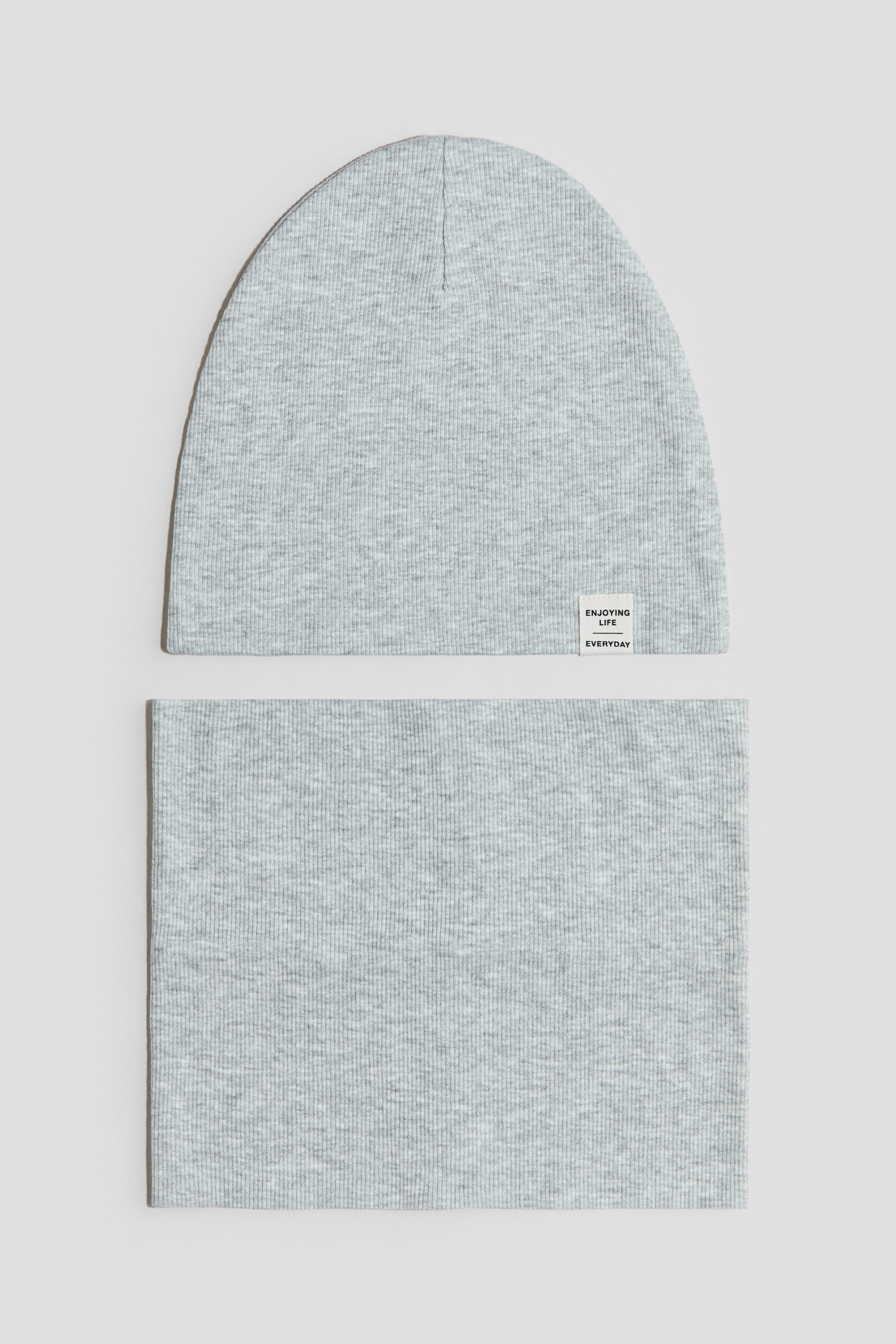 Größeres Bild ansehen: Ein hellgraues, geripptes, doppellagiges Baumwolljersey-Beanie und ein passender Schlauchschal liegen flach. Auf der Vorderseite des Beanies befindet sich ein kleines weißes Etikett auf der rechten Seite, das "enjoying life everyday" zeigt.
