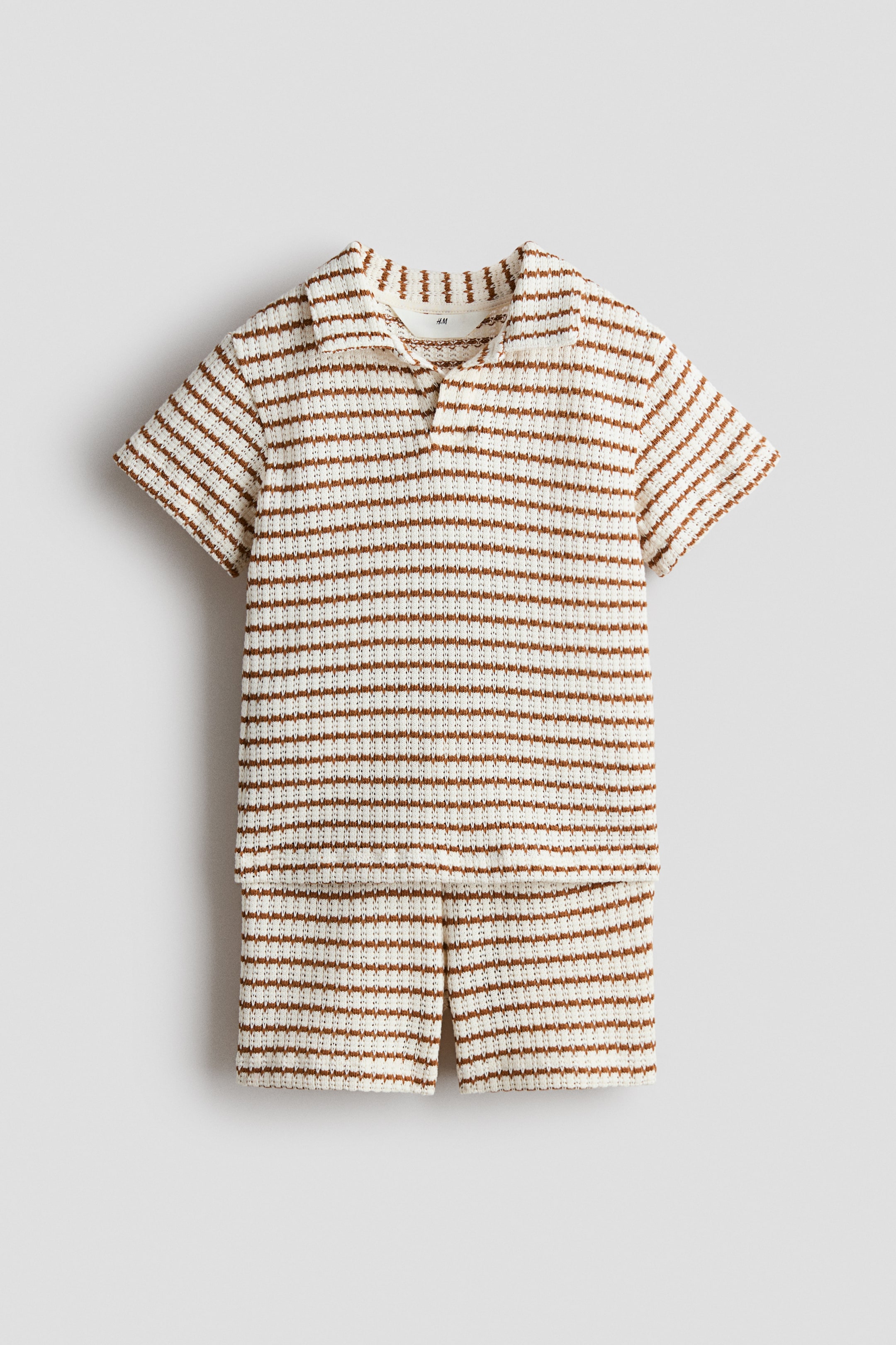 Grotere afbeelding bekijken: Een tweedelig breiset toont een kort mouwloos poloshirt en bijpassende shorts, beide met horizontale bruine strepen op een crèmekleurige achtergrond. Het shirt is ontworpen met een open kraag en een V-hals placket, en de shorts hebben kleine zijsplitjes aan de zoom.