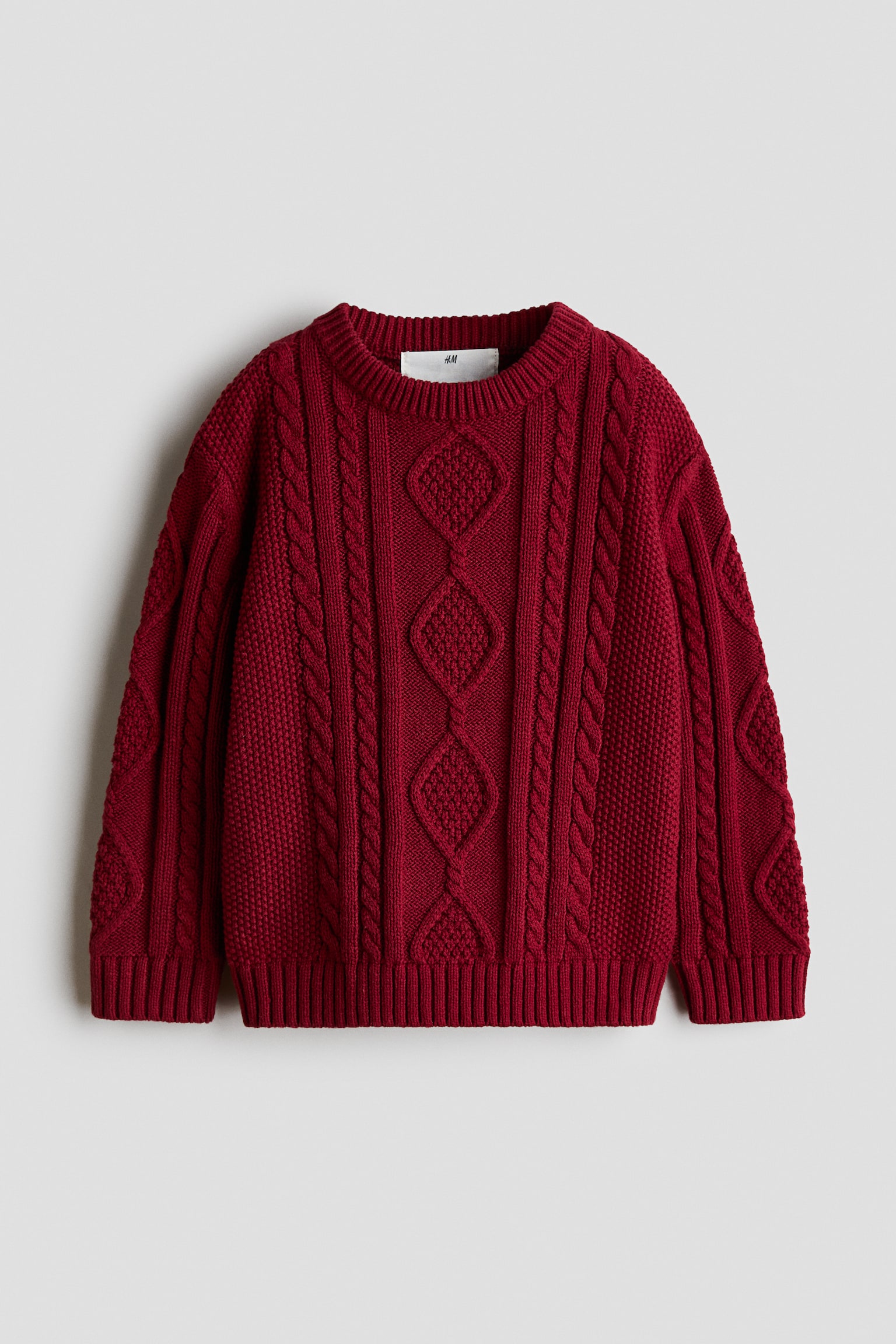 Un maglione a maglia intrecciata, dal taglio morbido e di un rosso intenso, è ripreso frontalmente e presenta un girocollo rifinito a coste, spalle scese e maniche lunghe. Il maglione mostra elaborati motivi verticali a trecce e a rombi su corpo e maniche, completati da polsini a coste e orlo a coste.