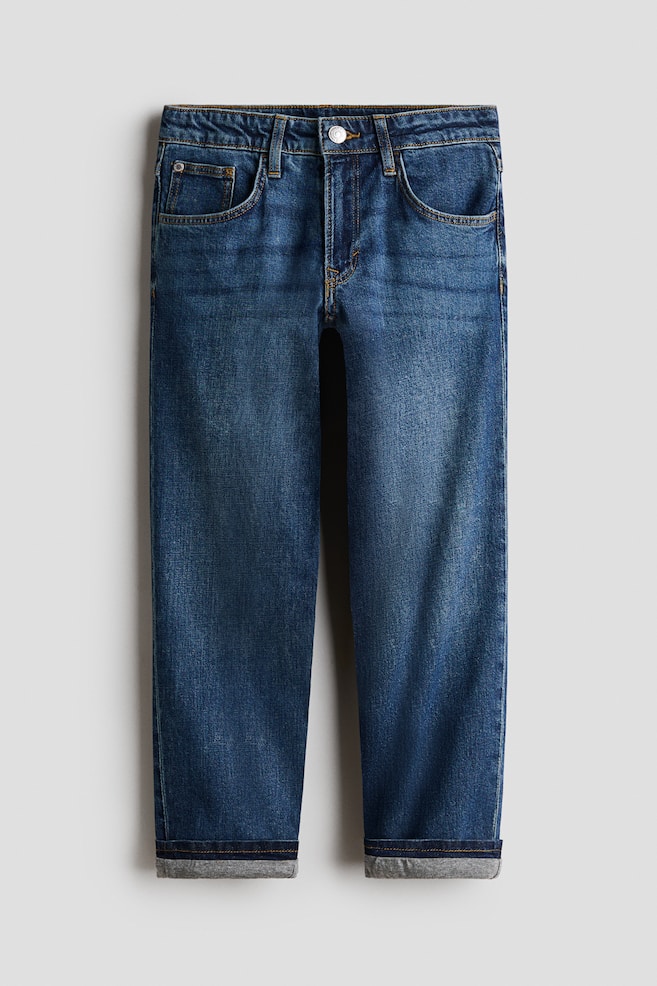 Blue jeans are shown in a front view, featuring classic five-pocket styling with contrast stitching, belt loops, and a metal button fastening at the waist. The straight-leg denim shows subtle fading along the thighs and is finished with turned-up hems revealing a grey lining. - 1