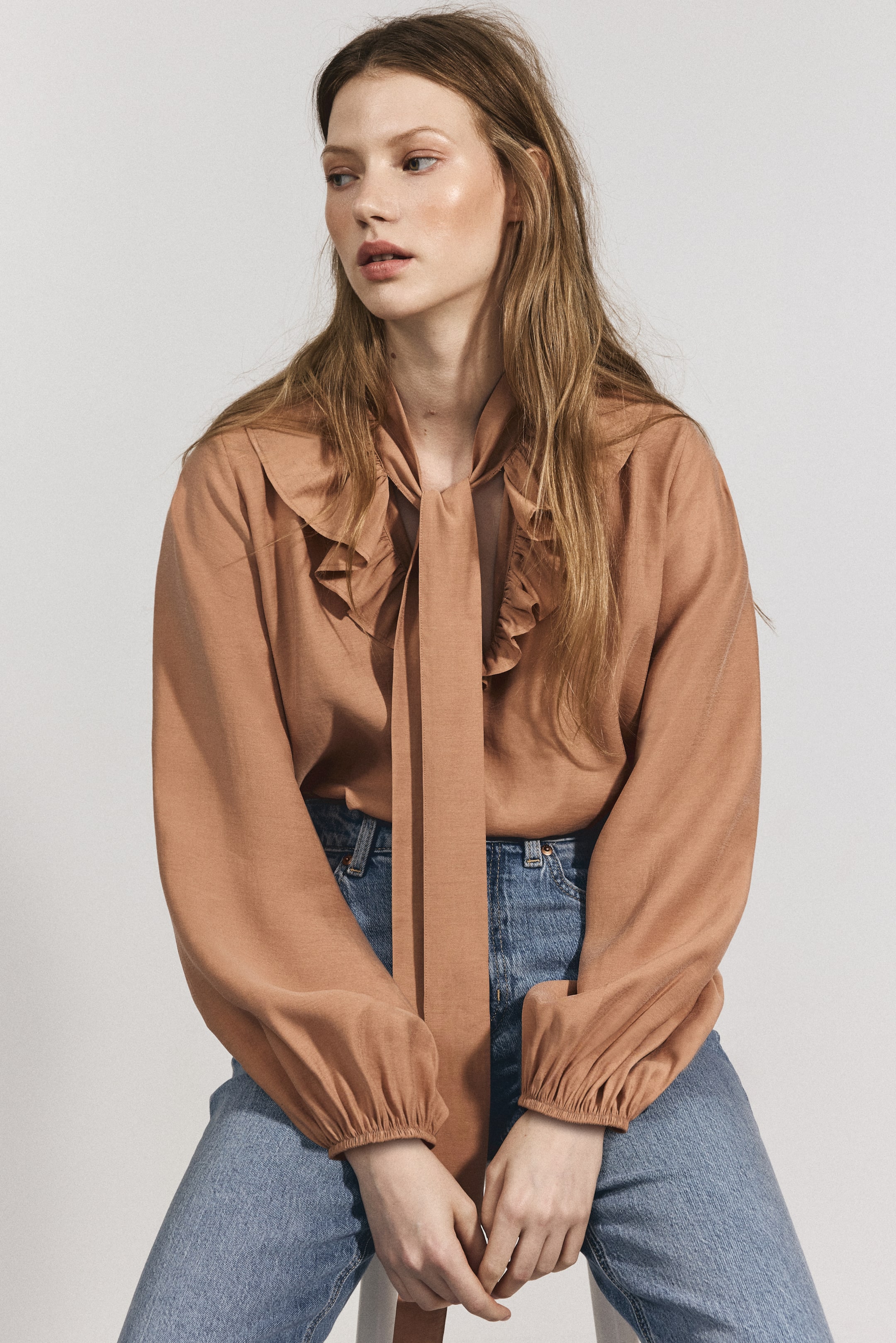 Büyük resmi görüntüle: Fırfırlı yakası ve büzgülü manşetli uzun kolları olan ten rengi bir fiyonk bluz, mavi kot pantolonun içine sokularak giyiliyor.