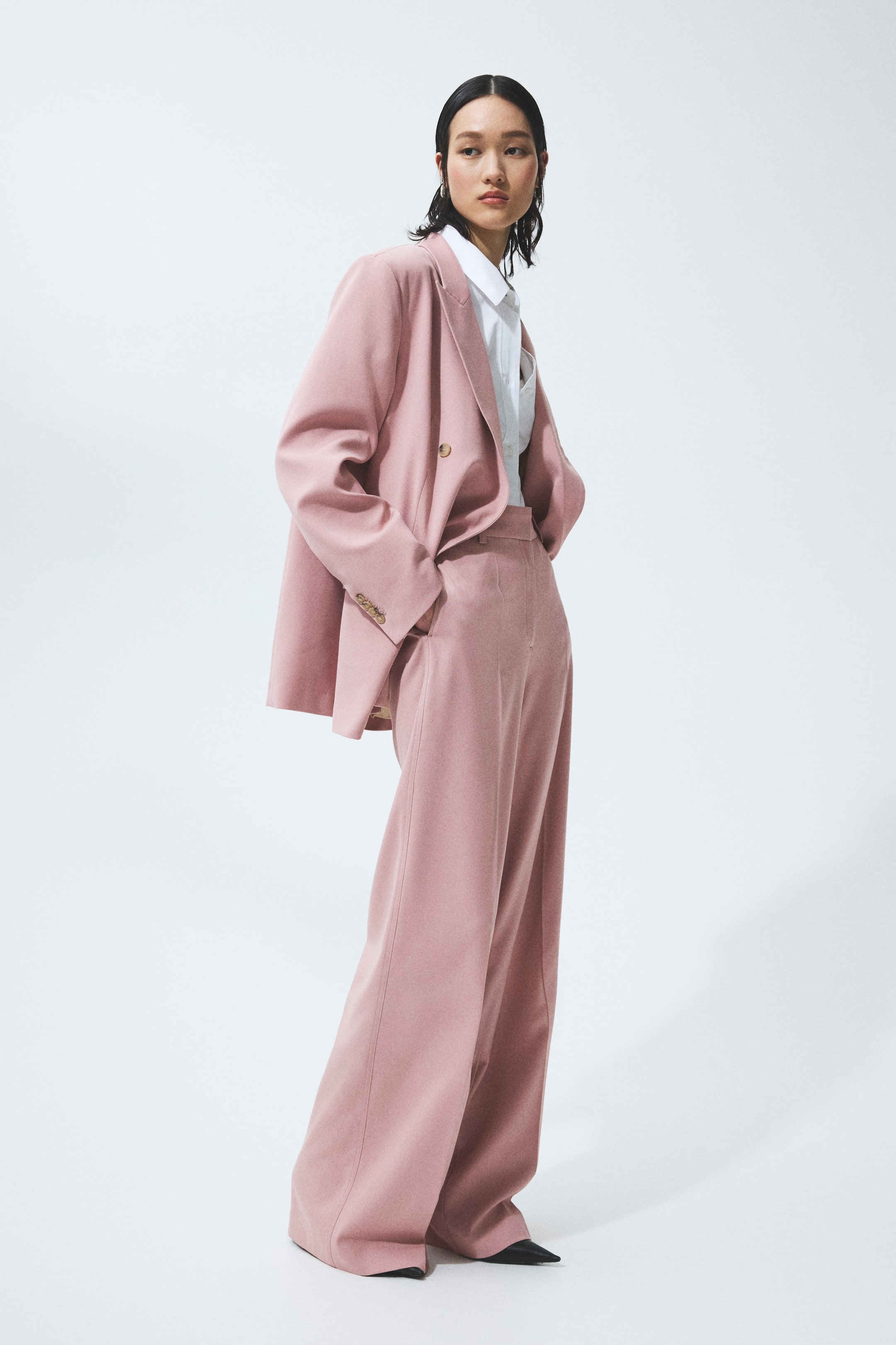 הצגת תמונה גדולה יותר: חליפת מכנסיים ורודה מוצגת מזווית של שלושה רבעים, המדגישה את הגזרה הרחבה והנינוחה שלה, וכוללת בלייזר כפול חזה עם דשים חתוכים, שני כפתורים קדמיים בצבע זהב ושלושה כפתורים קטנים יותר בצבע זהב על השרוולים, מעל חולצה לבנה עם צווארון. המכנסיים התואמים בגזרה גבוהה מעוצבים בגזרה רחבה, הנאספת בקרסוליים.